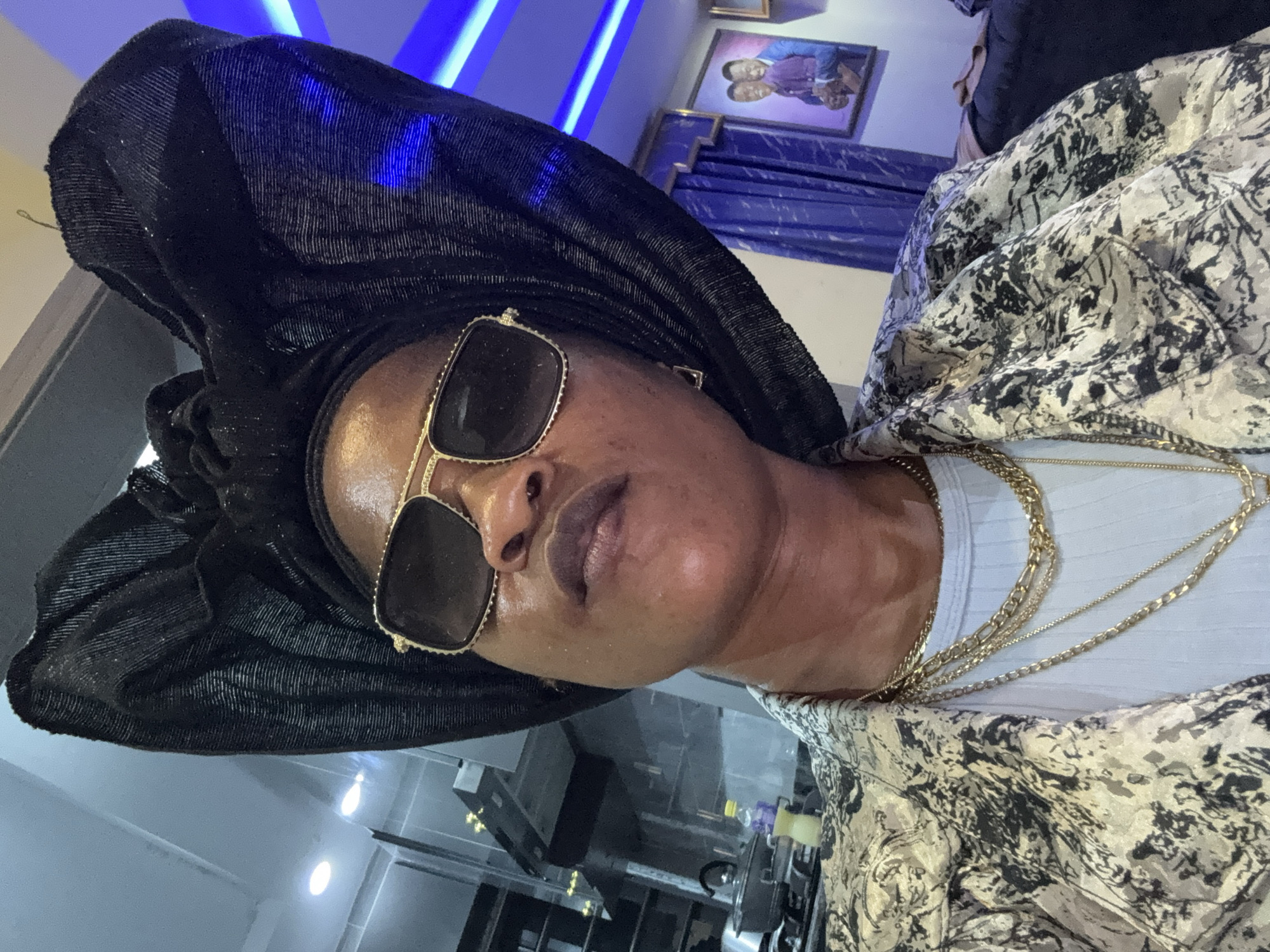 A medium shot captures a Black woman from the chest up, looking up and to her left. She is wearing a large, black headwrap and sunglasses with gold rims. Around her neck, she has several layered gold necklaces. She is also wearing a patterned jacket with a black and white abstract design over a light blue ribbed shirt. The background is blurred but suggests an indoor setting with kitchen appliances on the left, blue LED lighting strips on the right, and a framed portrait of two men on the wall. The lighting is artificial and casts a warm glow on the woman's skin. The overall impression is of a posed portrait, likely taken indoors. The location is identified as Ajara, Nigeria.