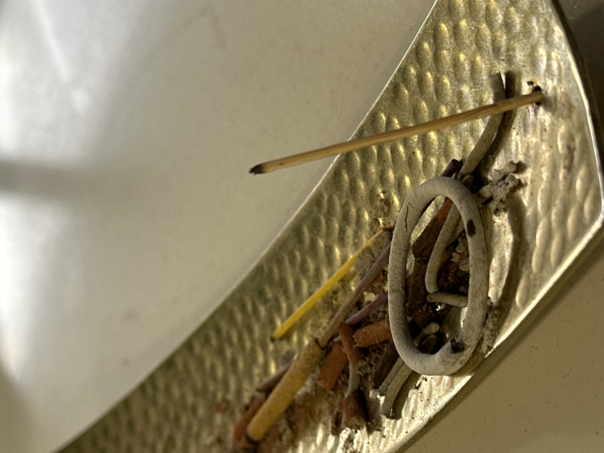 The image shows a close-up, top-down view of an ornate, gold-colored metal incense holder. The holder is long and rectangular with a textured, hammered surface. It contains several burnt-out incense sticks, broken pieces of incense, and a ring of ash. One unburnt incense stick stands upright in a hole on the right side of the holder, its tip smoldering with a faint wisp of smoke. The background is a blurred, light-colored surface, possibly a table or countertop. The scene is indoors, and no specific location in Nevinnomyssk, Russia can be identified from the image.