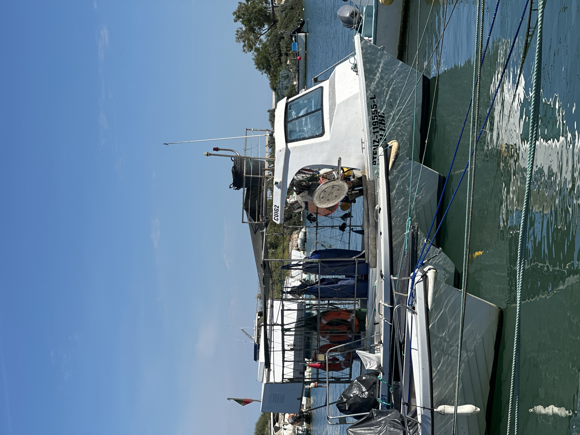 A daytime, medium-shot captures two boats moored side-by-side in the port of Olhão, Portugal. The sky is bright blue.
The foreground boat is white, with a rear portion covered by a black tarp. A red and green Portuguese flag is visible on the stern of the boat. Several orange life preservers are stacked on the rear of the boat.
The boat docked closer to the viewer features a white cabin and a large winch-like device on its deck. The hull of this boat is marked with "DTEIZ-119555-L EBE". Ropes are draped from the boat to the dock.
The backdrop consists of trees, buildings, and other vessels lining the waterfront of Olhão. The water surrounding the boats reflects the sky and surrounding structures.