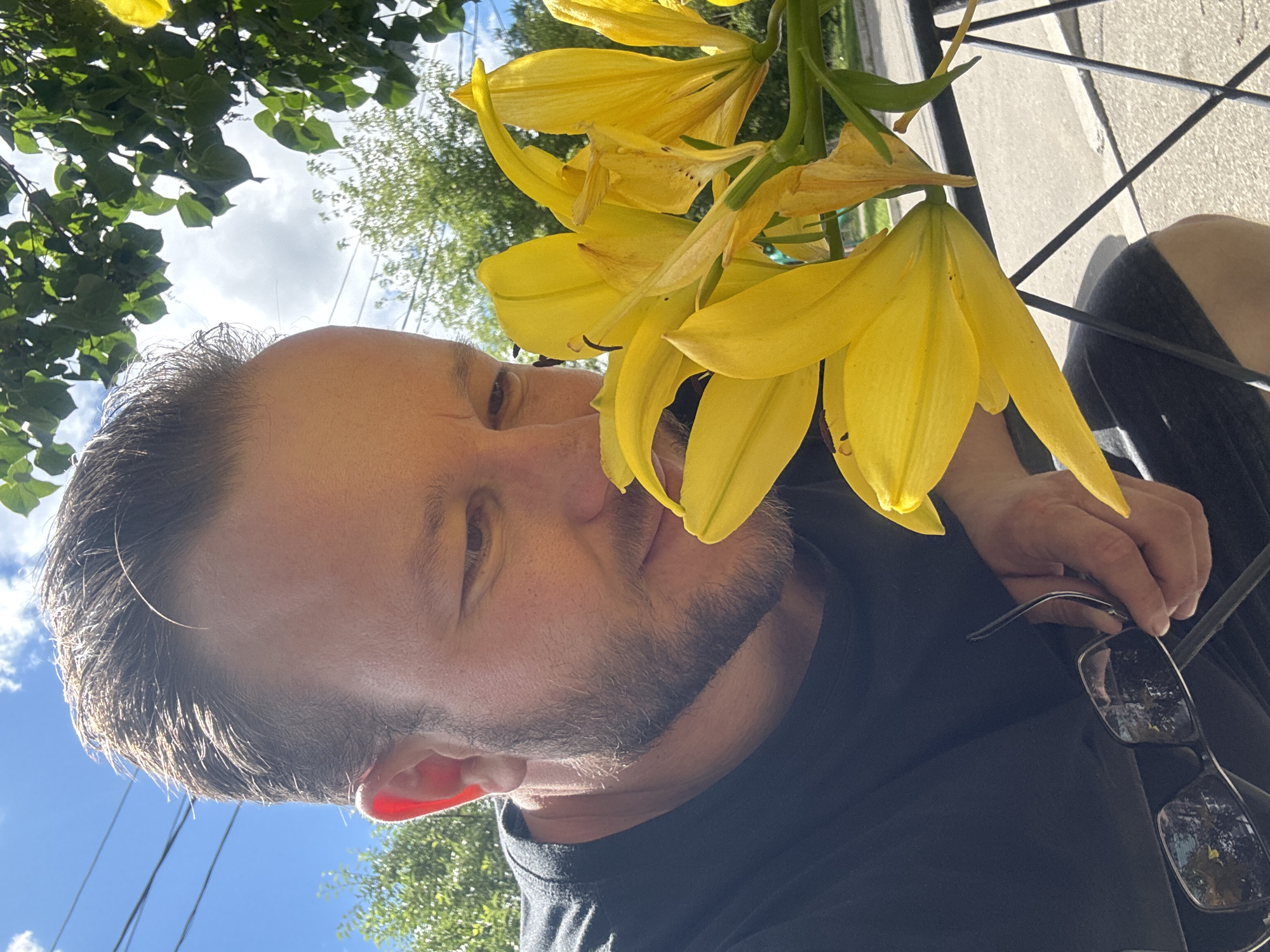 A man with short brown hair and a beard is shown from the chest up, looking down at a cluster of yellow lilies and smelling them. He is wearing a black t-shirt and holding a pair of glasses in his right hand. The background shows green trees, a blue sky with clouds, and utility wires, suggesting an outdoor setting in a city. The lilies are bright yellow and appear to be in bloom, with some petals slightly wilted. A metal fence or railing is visible in the foreground. The image does not provide any specific information to determine the location as Blagoveshchensk, Russia.