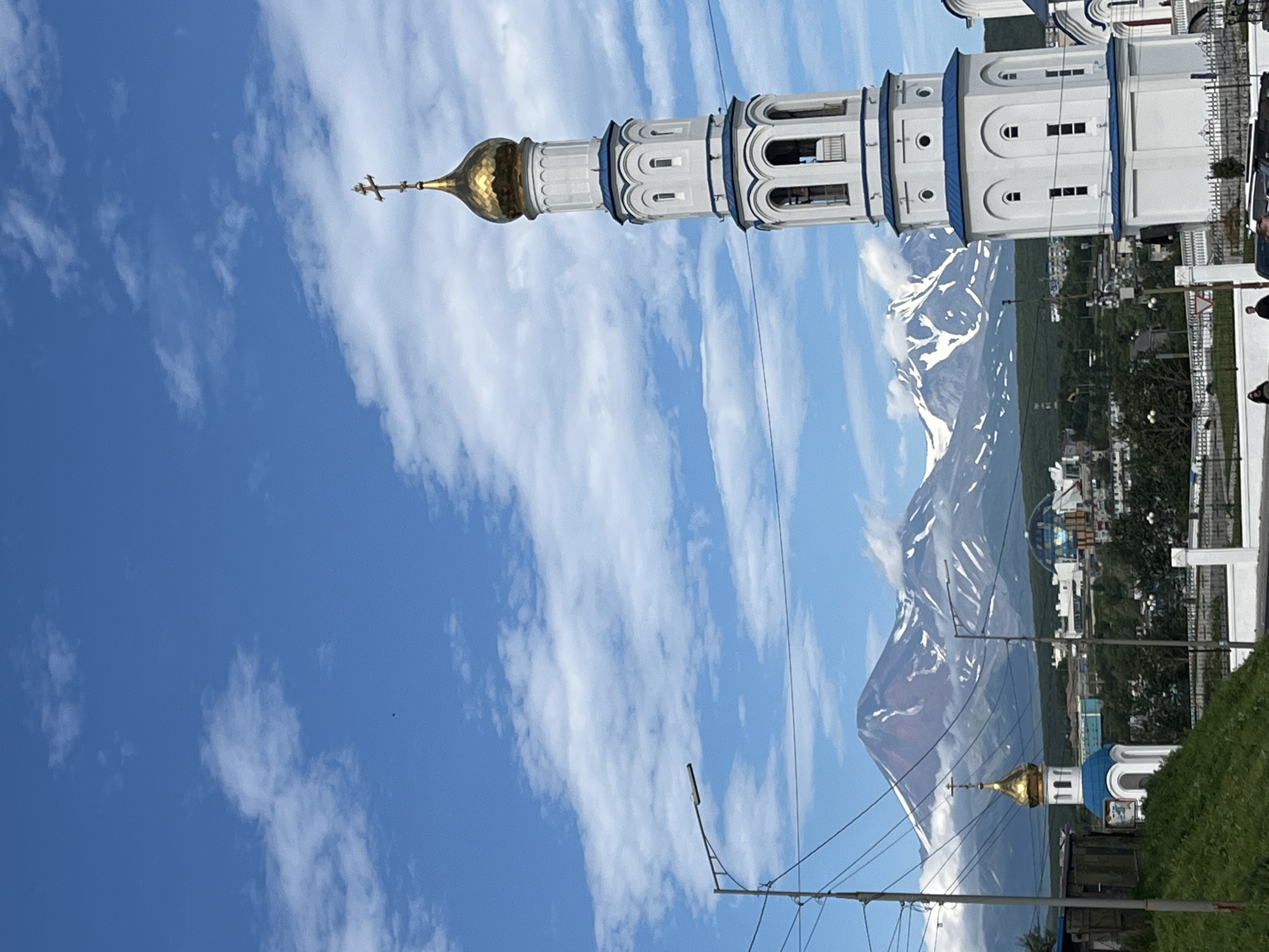 A tall, white, multi-tiered bell tower with a golden dome and a cross stands on the right side of the frame. It has blue trim and arched windows. In the middle ground, a smaller church with a blue dome is visible, also adorned with a golden cross. Behind the churches, a large, snow-capped volcano dominates the background, its peak partially obscured by clouds. The sky is bright blue with scattered white clouds. Several utility poles and wires are present in the foreground and middle ground. The bottom of the image shows a grassy hill and a fence. Two individuals are barely visible in the lower right corner. A large domed building is also visible in the middle distance. The scene appears to be set in the city of Petropavlovsk-Kamchatsky, Russia, given the presence of the volcano Klyuchevskaya Sopka.