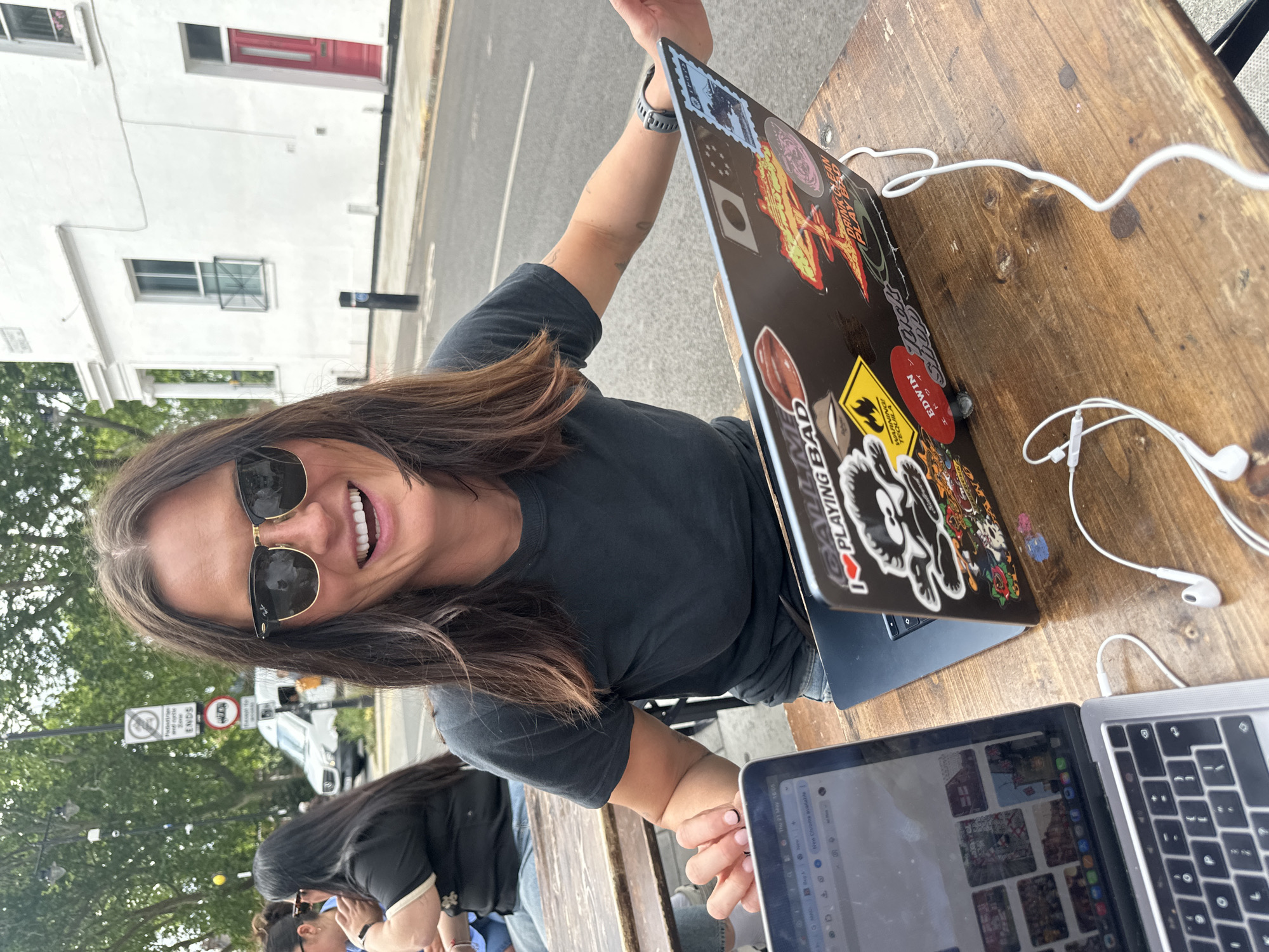 The image shows a woman wearing sunglasses and smiling at the camera. She is sitting at an outdoor wooden table with two laptops on it. One laptop is open and showing images, while the other has stickers on its cover. White earphones are also on the table. The woman is wearing a dark t-shirt. In the background, other people are sitting at a table. A street scene is visible with cars and buildings. Road signage indicates the image was taken in the city of London, United Kingdom.