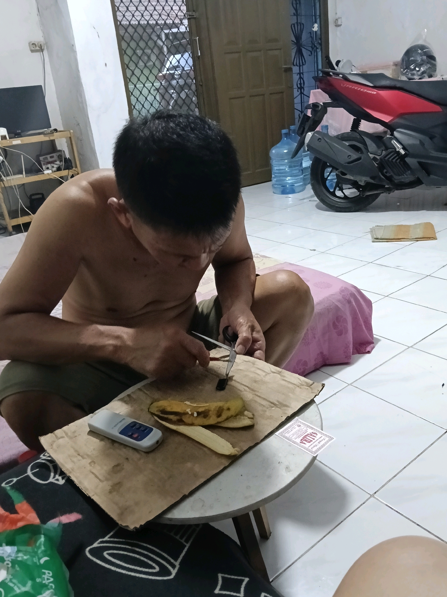 An unclothed person is squatting on the floor in Jatimulya, Indonesia, preparing a banana with scissors. The person is seated on a pink cloth-covered object, possibly a stool or cushion, with a cardboard piece and a small silver device on it. The banana appears to be cut and partially peeled. To the right of the scene, a red and black motorcycle is parked. In the background, there are two large blue water jugs and a doorway with a barred window. The floor is tiled white.