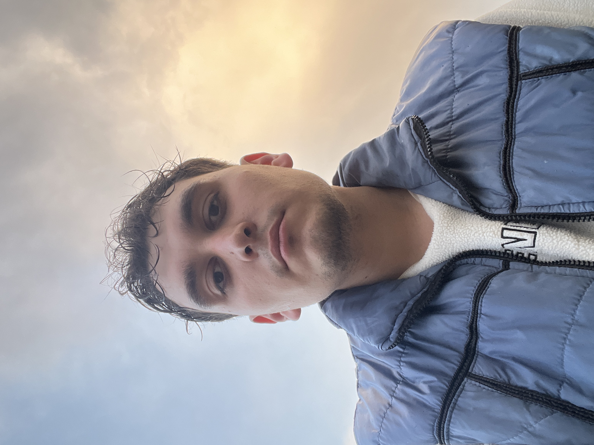 A young man with dark hair and a stubble beard is facing the camera, looking down. He is wearing a light blue puffer vest over a white fleece jacket. The background shows a cloudy sky with a patch of sunlight. The location is unknown.