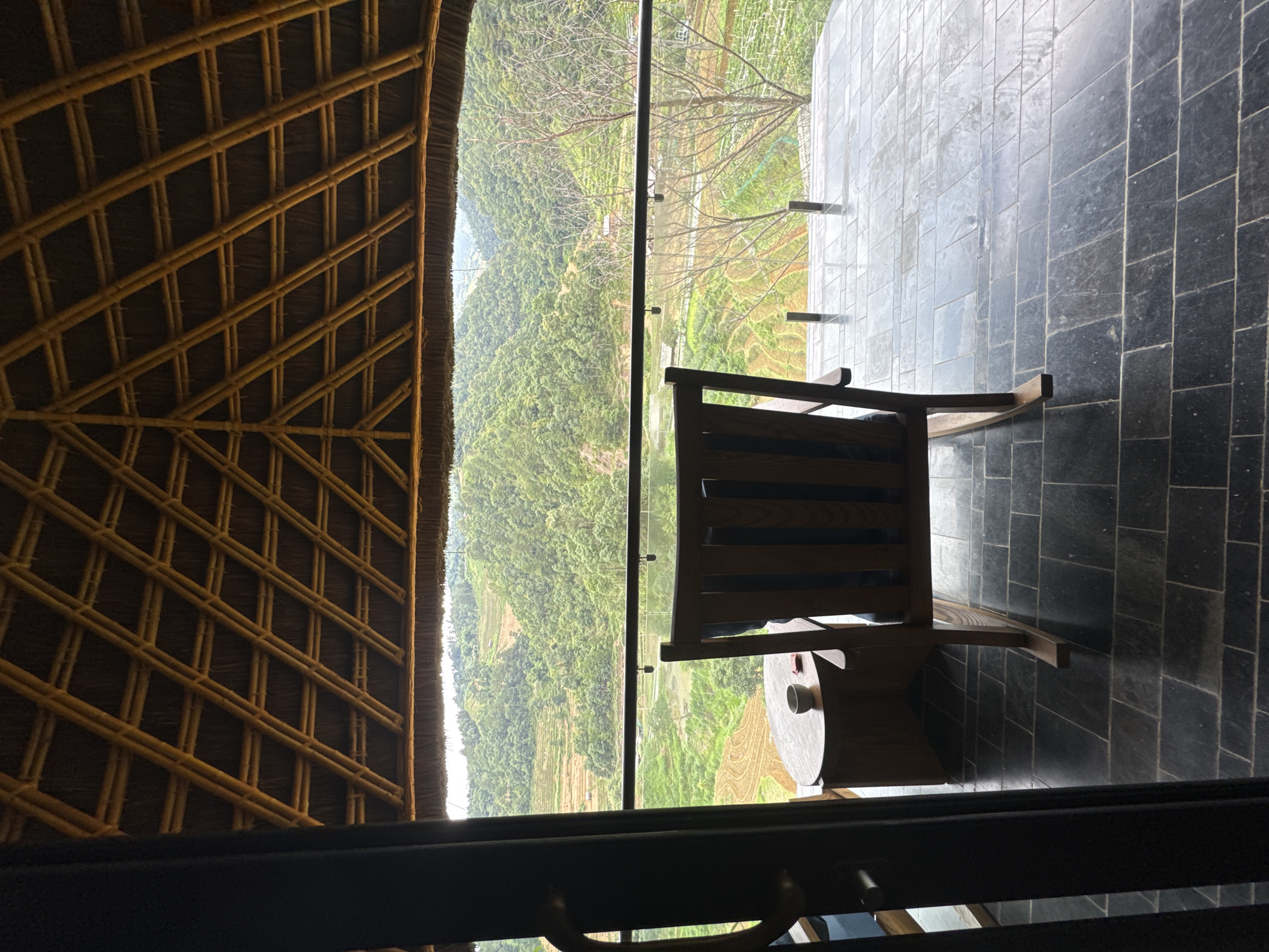 This image captures a serene outdoor balcony scene in Mù Cang Chải, Vietnam, offering a panoramic view of lush, terraced mountains. The vantage point is from inside a structure, looking out through a dark doorway onto a covered patio. Above, a striking, light-brown bamboo-framed roof with a thatched or woven material ceiling forms a precise grid pattern. The patio floor is paved with dark gray, rectangular stone tiles. A wooden rocking chair with blue cushions is positioned on the patio, facing away from the camera and towards the expansive landscape. Adjacent to the chair is a small, round wooden side table holding a light-colored bowl and a small red object. A clear glass railing with dark support posts provides an unobstructed view of the exterior. Beyond the railing, the landscape unfolds with numerous verdant, forested mountains and hills. Distinctive rice terraces, some appearing flooded and reflective, others showing dry, patterned earth, are carved into the mountain slopes. In the far distance, a valley floor is visible under an overcast sky, casting a soft, diffused light across the scene. A few bare-branched trees stand on the right side of the view. No people are visible.