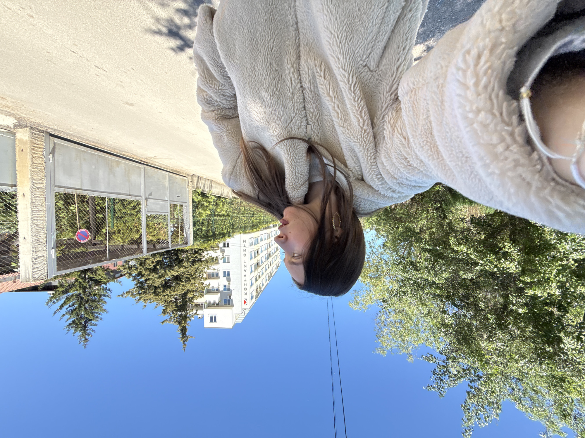 The image depicts a woman wearing a light-colored, fluffy jacket. Her head is oriented towards the upper left corner of the image. In the background, there's a blue sky with green trees and a tall, white building with multiple windows, as well as a metal gate. This location is in Sofia, Bulgaria.