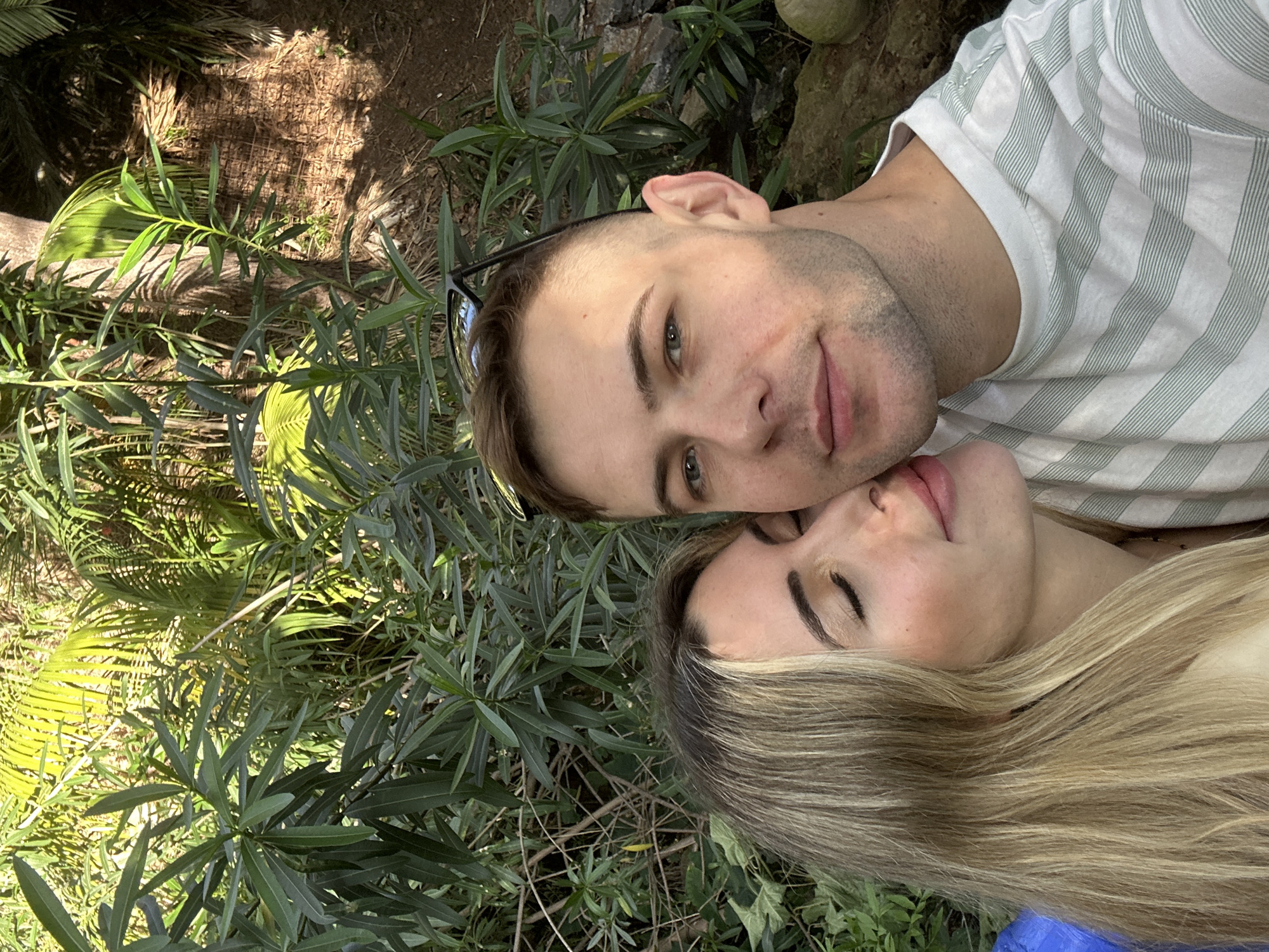 A man and a woman are lying together in what appears to be a garden or wooded area. The man has short brown hair and blue eyes, and he is wearing a striped t-shirt. The woman has long blonde hair and is lying with her eyes closed. They are surrounded by green foliage, including plants with long, narrow leaves and a palm-like tree in the background. It is difficult to determine the exact location of the photo in Altea, Spain without further information, but based on the vegetation, it is likely a botanical garden or park in the area.