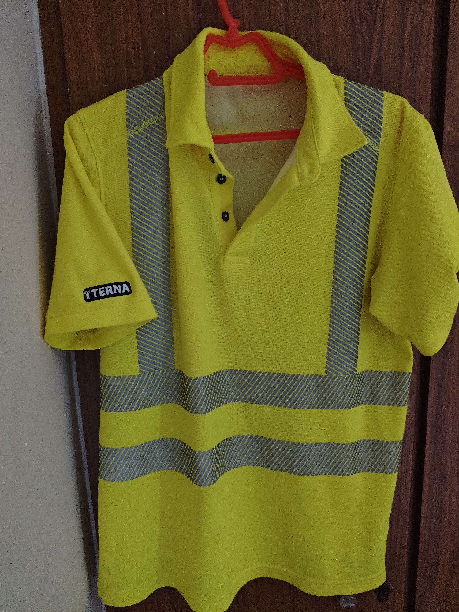 A bright yellow short-sleeved polo shirt with reflective silver stripes is hanging on a red plastic hanger. The shirt has a collar and a three-button placket. A black logo with white lettering that reads "TERNA" is visible on the left sleeve. The shirt is hanging against a background of a light-colored wall on the left and a dark wooden door on the right. The image appears to have been taken indoors, likely in a residential or storage setting. There is no discernible connection to the city of Athens, Greece.