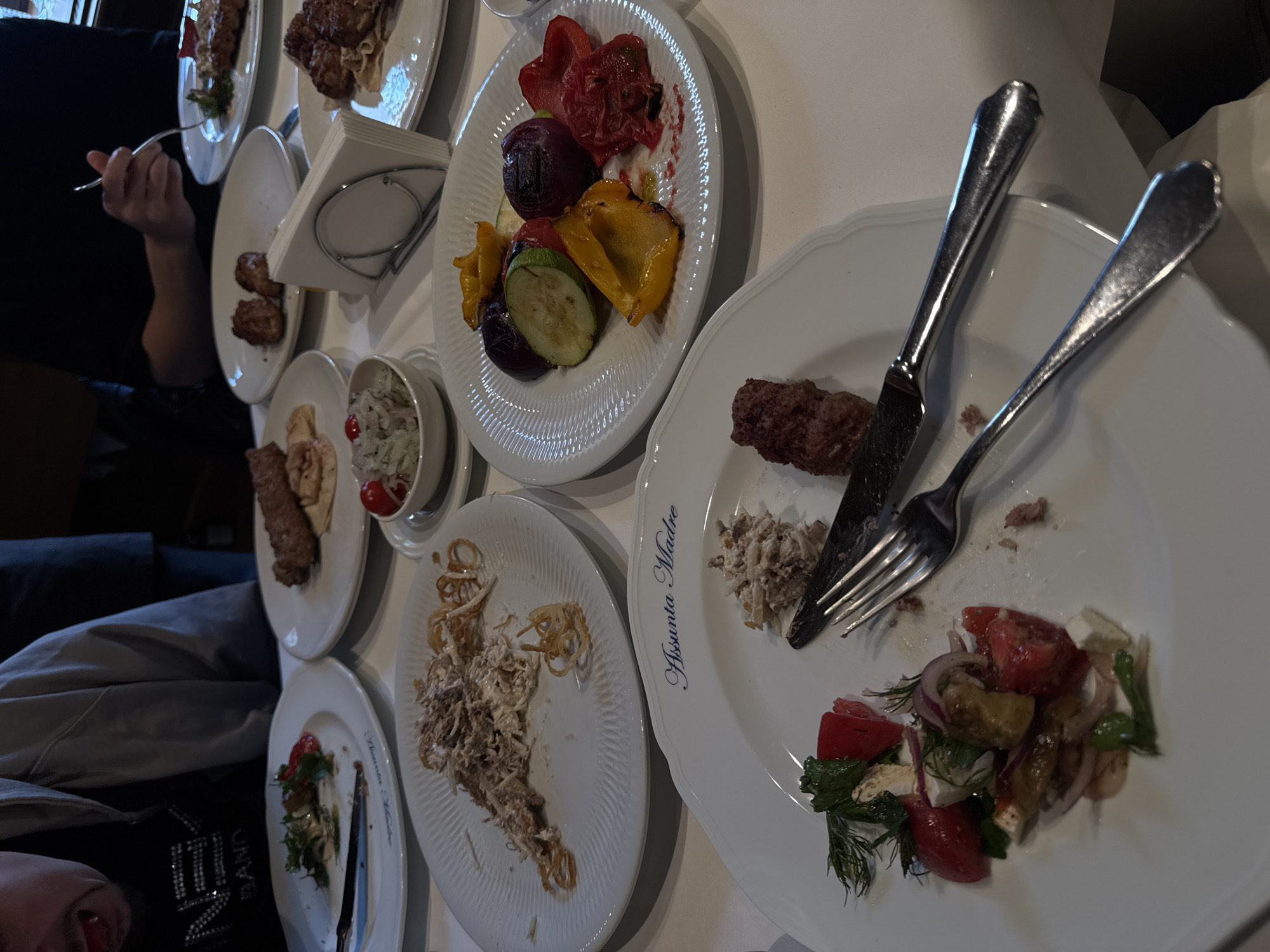 The image shows a dining table laden with plates of food, cutlery, and napkins. Dishes include grilled vegetables, salads, and meat skewers. Some plates are partially consumed.
One plate reads "Assunta Madre."
The general presentation is that of a shared meal being enjoyed. The location is likely within a restaurant in Moscow, Russia, due to the name 'Assunta Madre' which is known to be a branch of a restaurant with locations in Moscow.