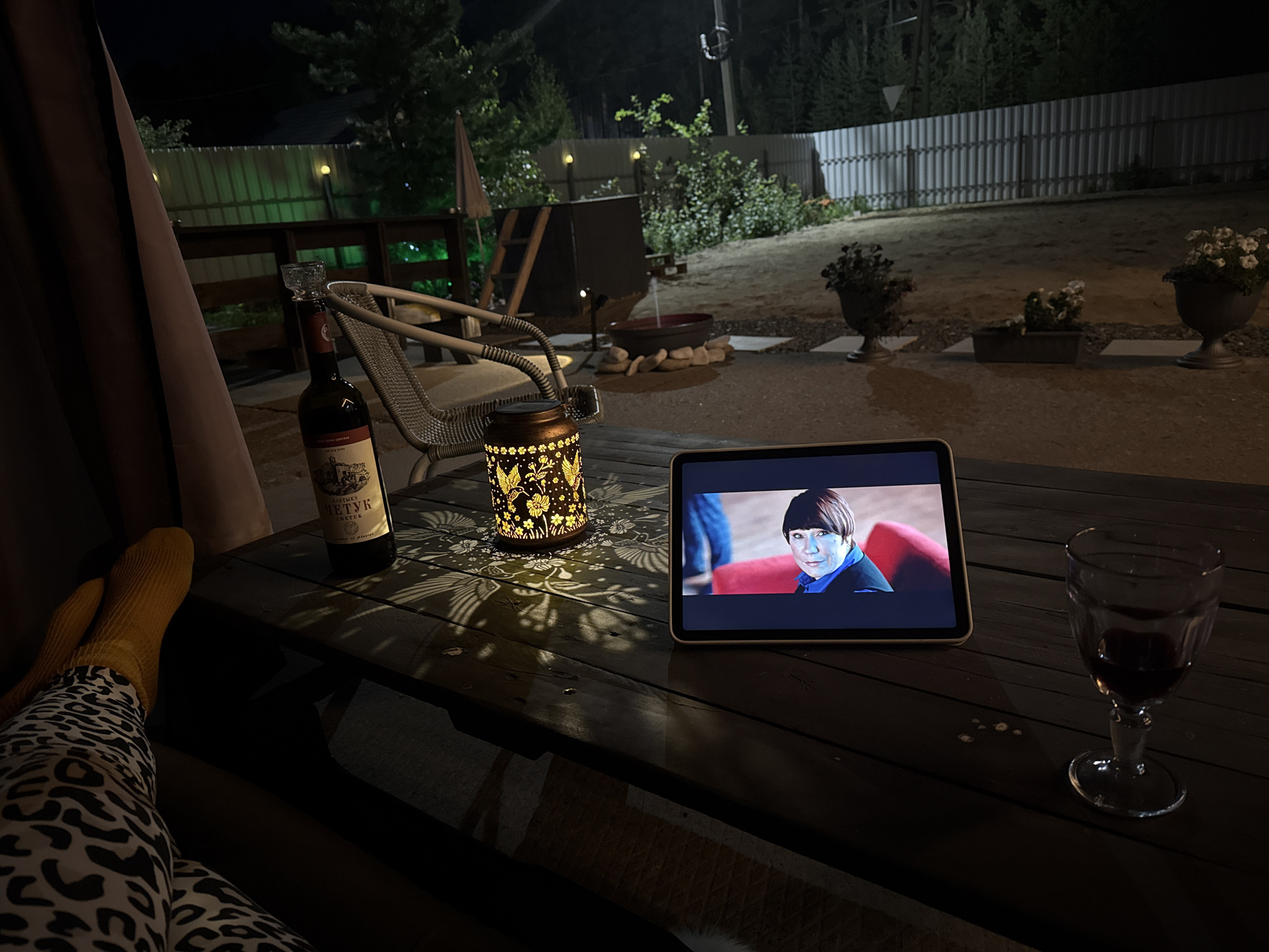The image depicts a dark outdoor scene, likely a backyard patio at night, in Sovetskiy, Russia. A person's legs, clad in patterned leggings and yellow socks, are visible in the foreground on the left, suggesting they are seated at a wooden table. On the table, there is a bottle of red wine, a decorative lantern casting patterned shadows, a tablet displaying an image of a person with short brown hair and a blue shirt, and a wine glass filled with red liquid. Behind the table, a wicker chair is visible. The background shows a fenced yard with greenery, trees, and outdoor lighting. The overall ambiance is one of relaxation and solitary entertainment.