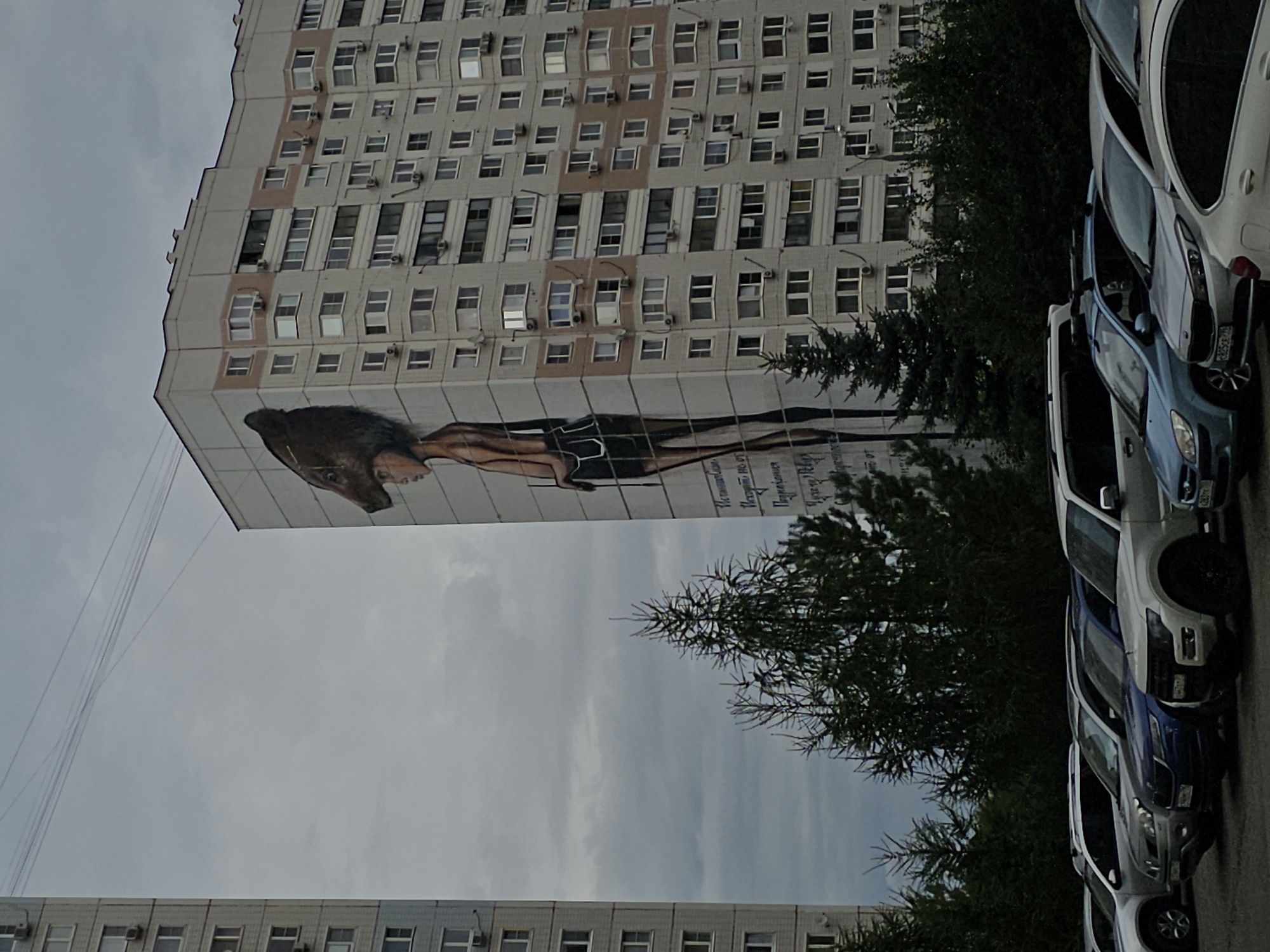 The image depicts a mural of a person with a large, dark, animal-like head covering their own on the side of a multi-story apartment building in Odintsovo, Russia. The building has numerous windows and air conditioning units. In the foreground, several parked cars are visible, including SUVs and sedans. Evergreen trees form a line between the building and the parking lot. The sky is overcast and appears to be cloudy. The mural occupies a significant portion of the building's facade.