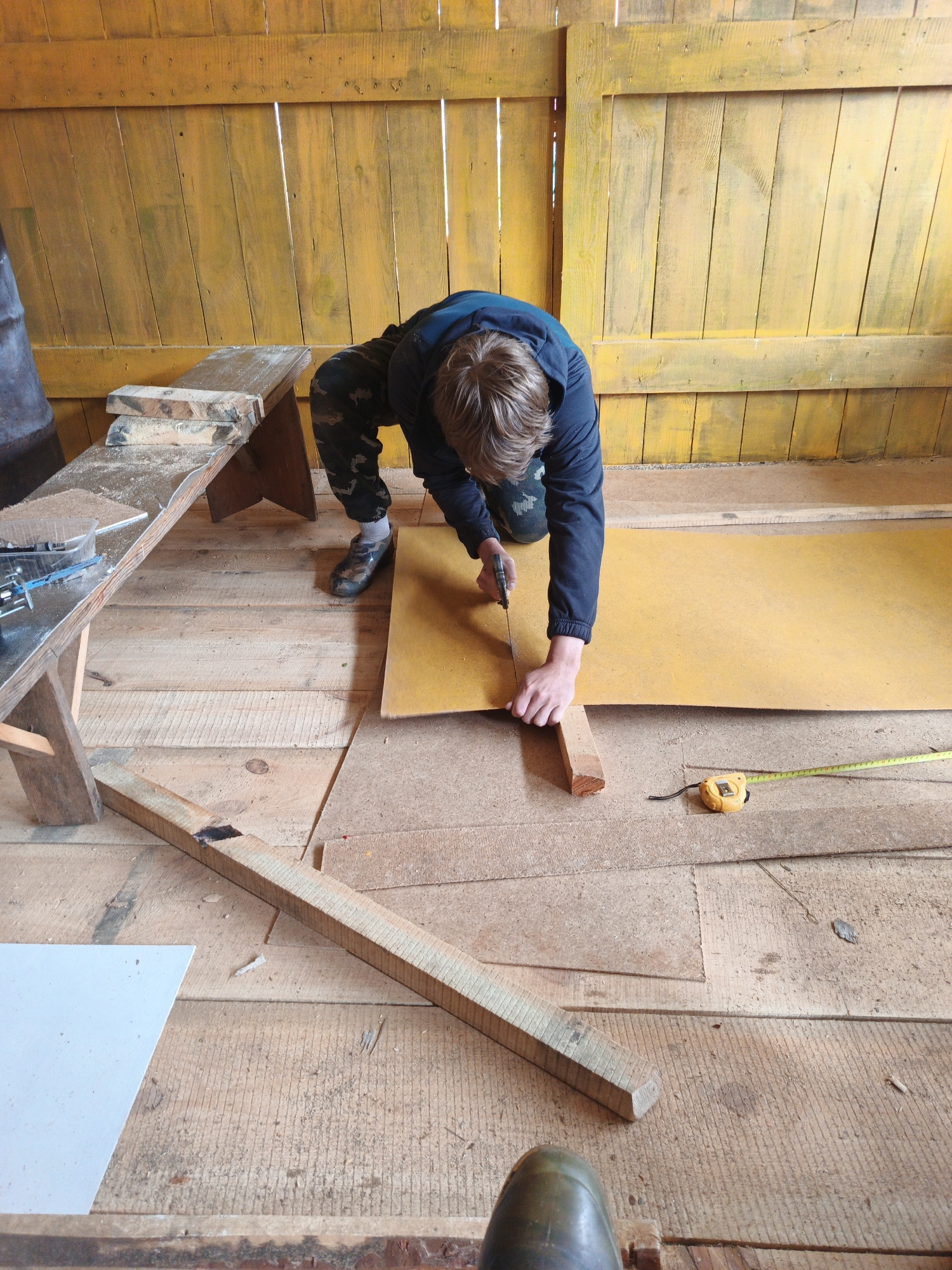 In a rustic wooden structure, possibly a workshop or shed, a young person with light brown hair, wearing a dark blue hooded jacket and camouflage pants, kneels on the floor. They are focused on cutting a large sheet of yellowish material, likely a flooring or insulation material, with a utility knife. A small wooden block is placed under the material to aid in the cut, and a measuring tape is visible to the right. The floor is made of rough wooden planks, and the walls are constructed from vertical yellow-painted wooden boards. The environment appears to be a construction or renovation site, with some lumber and tools scattered around. The scene does not provide any specific indicators to place it in Islambaevo, Russia.