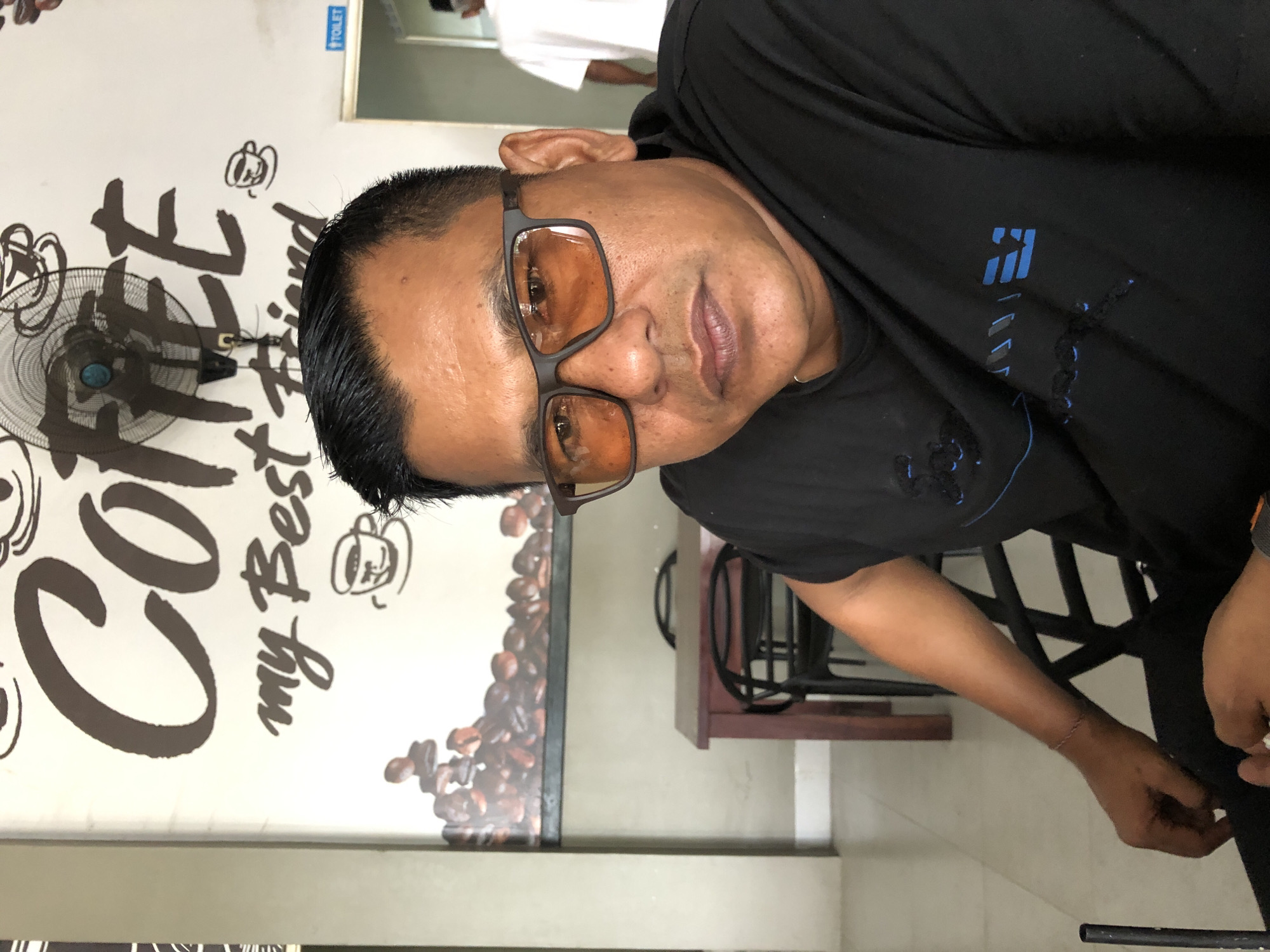 A medium shot photograph captures a man wearing a black t-shirt and glasses, seated in what appears to be a cafe. The background features a wall adorned with a large coffee-themed mural depicting coffee beans and the words "Coffee my Best Friend." A ceiling fan is mounted above the mural. To the right of the frame, a doorway is visible with a sign indicating "TOILET." In the mid-ground, a wooden table with chairs is set up. The man is looking directly at the camera, with a neutral expression. The lighting suggests an indoor setting, likely during the daytime. The location is identified as Geulumpangsulu Timu, Indonesia.