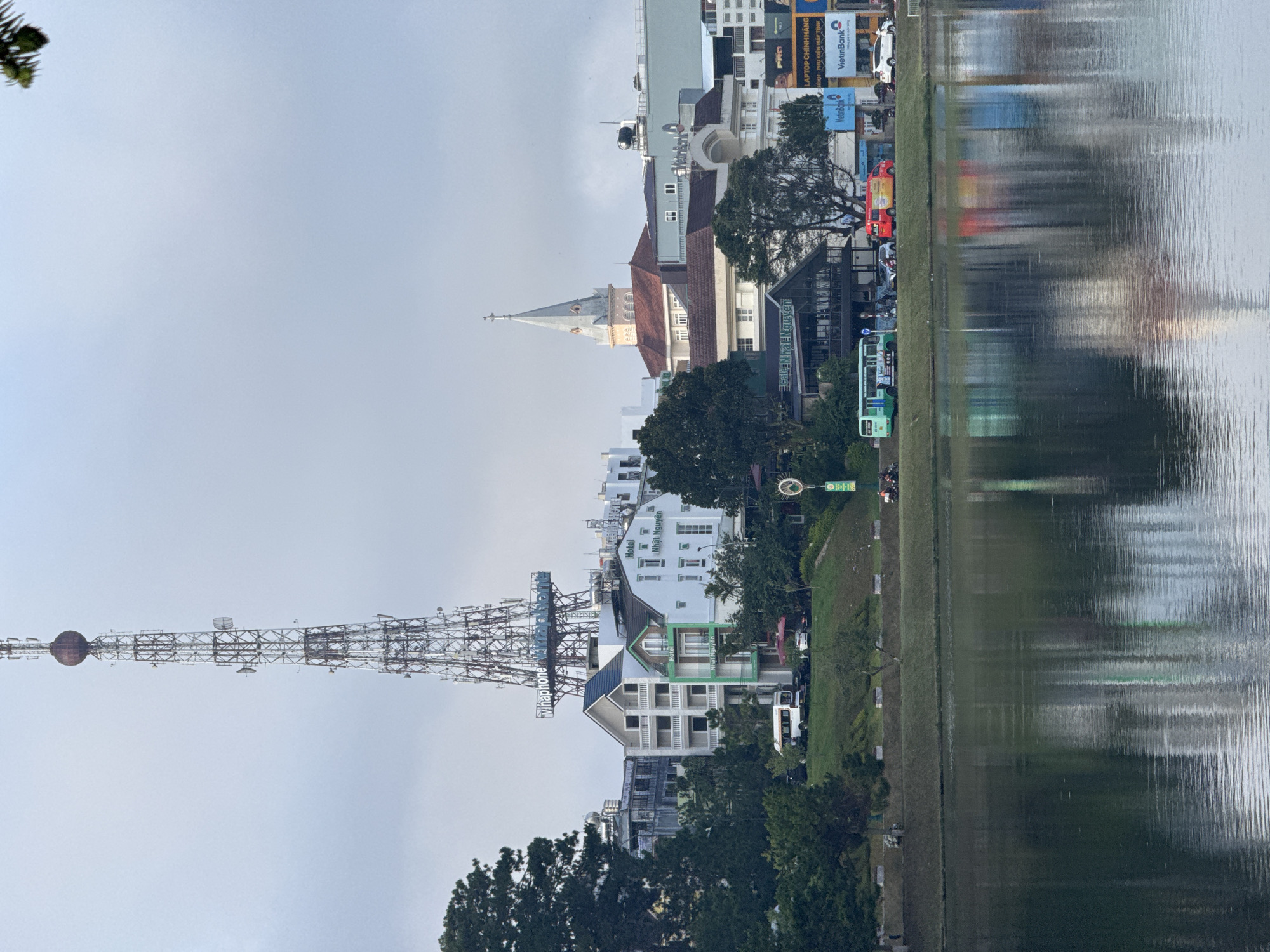 The image depicts a tranquil lakeside scene in Da Lat, Vietnam, likely taken from the Xuan Huong Lake area, a popular tourist destination. In the foreground, the calm surface of the lake reflects the overcast sky and the buildings lining its shore. A prominent landmark, a tall, lattice tower with a spherical structure at its apex, dominates the skyline. This tower is likely a telecommunications tower, possibly belonging to Vinaphone, as indicated by signage.
Behind the tower, various buildings are visible. A light-colored hotel with green accents, labeled "Hotel Nhat Nguyen," stands out amongst the others. Further back, a church with a distinctive spire adds architectural interest. The shoreline is lined with trees and a grassy embankment, with a road running parallel to the lake. Several vehicles, including a white bus, a red van, and cars, are parked or moving along the road. The overall atmosphere is subdued due to the cloudy weather, but the scene suggests a typical day in this scenic Vietnamese city.