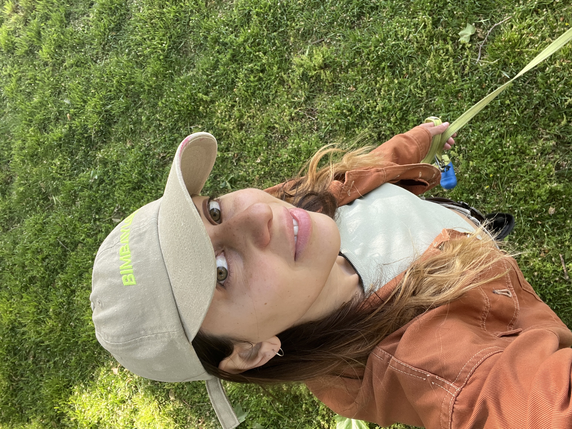 A low-angle selfie captures a young woman outdoors against a backdrop of lush green grass. The image is oriented such that the woman's head is tilted to the left, looking directly into the camera with wide, engaging green/hazel eyes. She has light skin with visible freckles and wears a small silver hoop earring in her left ear. Her lips are a natural pink, parted slightly to reveal a hint of white teeth.
She is wearing a light beige or tan baseball cap with "BIMBAY" visible in bright neon green text on the front. Her long, wavy hair, a mix of brown and blonde tones, falls around her face and shoulders. She is dressed in a light mint green top or t-shirt, layered under a rust-colored or terracotta-toned jacket with contrasting white stitching.
In her right hand, she holds a bright neon yellow-green leash, with a small blue object, likely a poop bag dispenser, attached near the handle. The absence of a dog suggests she might be about to walk one, or has just finished. The setting appears to be a sunny park, field, or lawn, characteristic of a pleasant day in an outdoor environment such as Barcelona, Spain. The bright, natural lighting indicates it is daytime.
