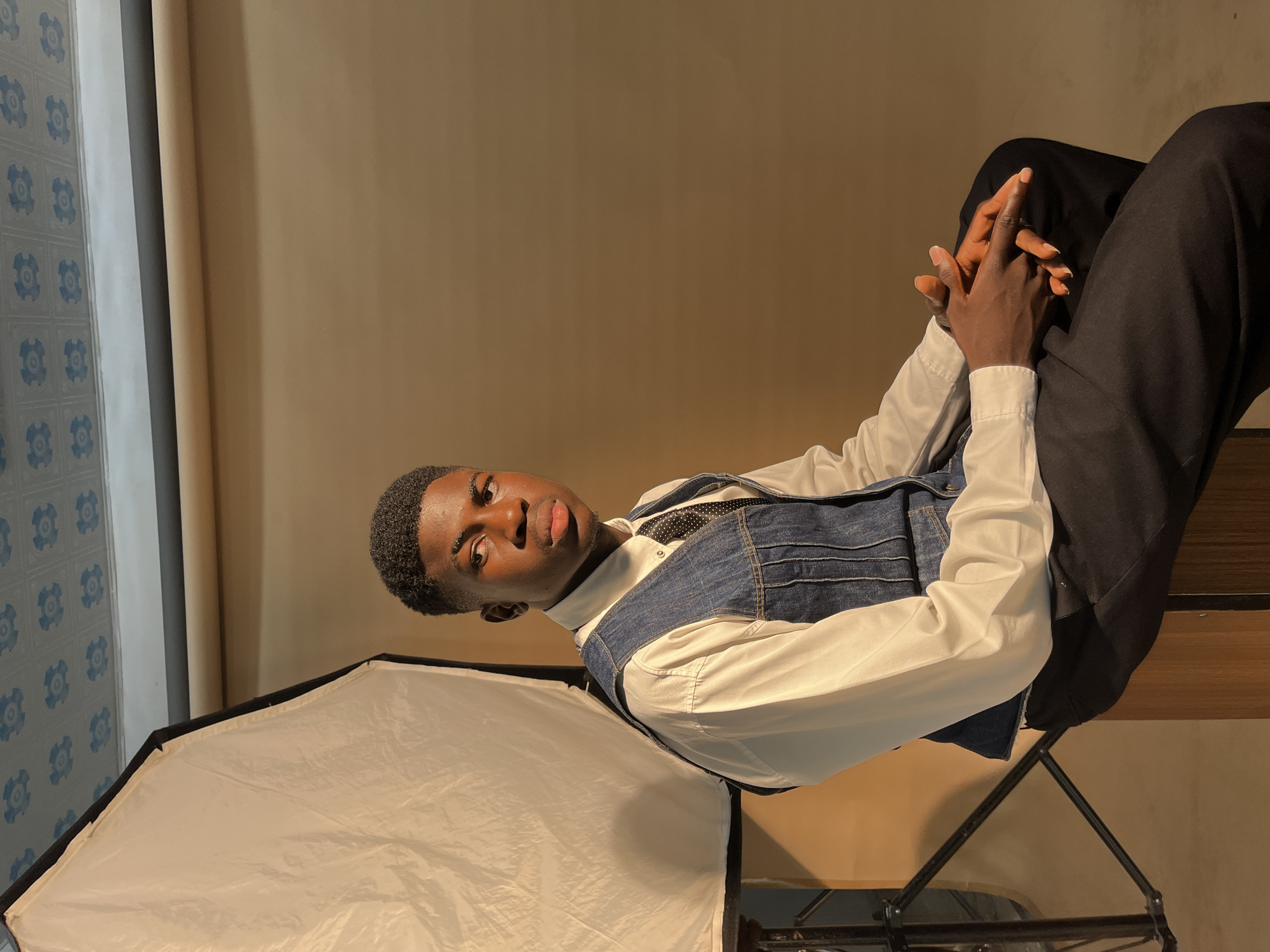 A young man is sitting in a chair in a room of Unknown, Unknown. He is wearing a white shirt, a denim vest, and black pants. His hands are clasped together. There is a softbox to his left and patterned wall to the left. The room is lit by a warm light.