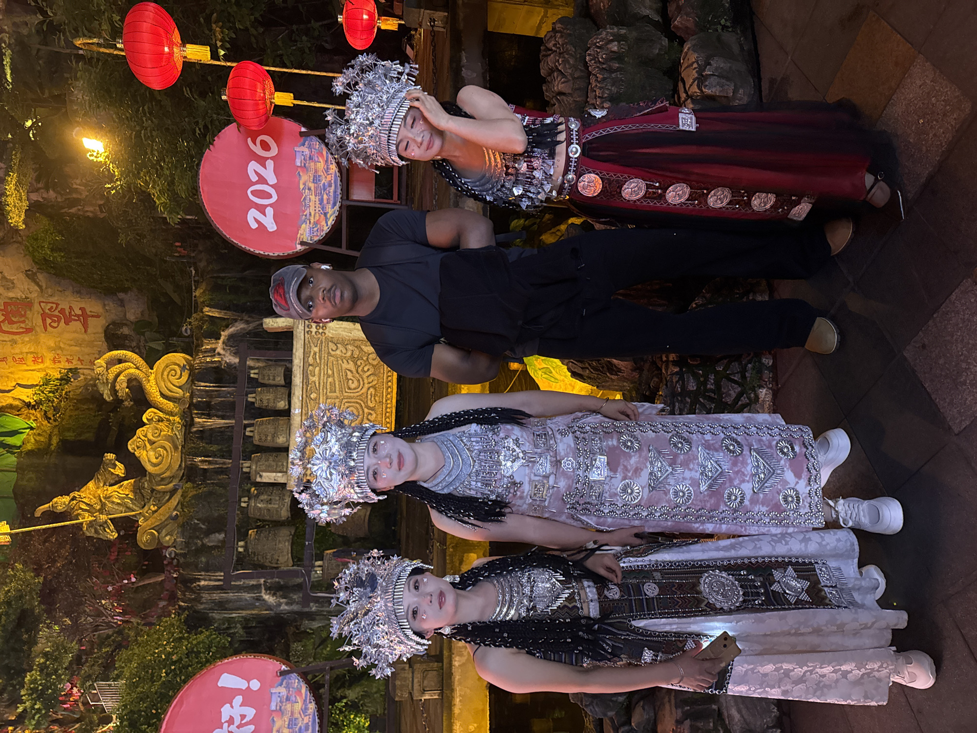 The image shows a man posing with three women dressed in traditional attire in what appears to be a tourist area in Yuzhong District, China. The man is in casual clothing, a black shirt and pants, and a cap. The women are wearing ornate silver headpieces and traditional costumes with intricate silver details. Red lanterns and a round sign with "2026" are visible in the background. Decorative golden statues and carved walls suggest a historic or cultural site. The lighting indicates that it is evening or nighttime. One of the women is holding a smartphone. The setting appears to be outdoors.