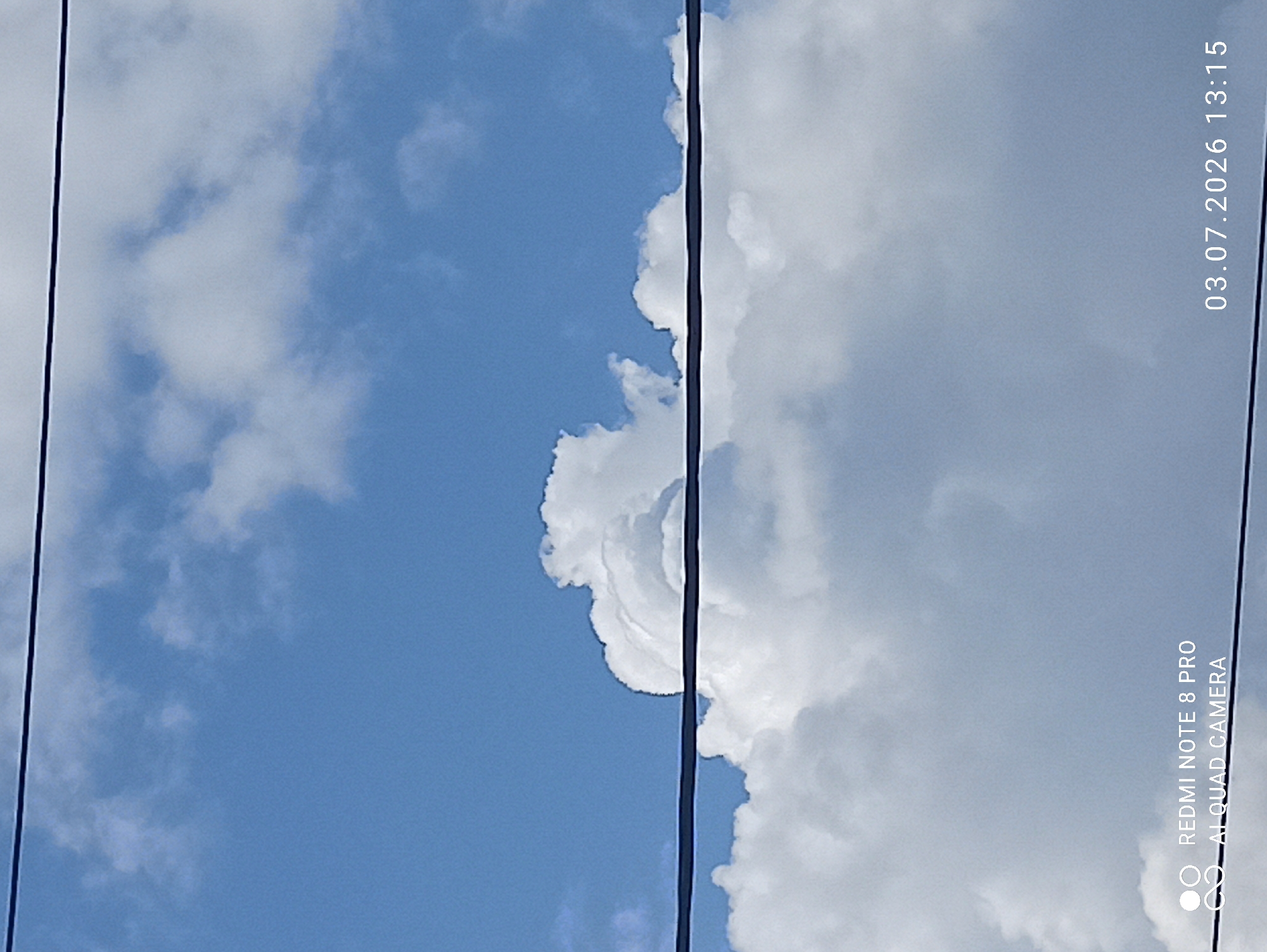 The image depicts a clear blue sky with scattered white, fluffy clouds. Two black wires traverse the frame horizontally, suggesting an outdoor, possibly urban or suburban, setting. The lower portion of the image shows more dense clouds. In the bottom left corner, a watermark reads "REDMI NOTE 8 PRO AI QUAD CAMERA". In the bottom right corner, a date and time stamp indicates "03.07.2026 13:15". The scene is devoid of people or any specific landmarks that would pinpoint a location within the city of Sovetskiy, Russia.