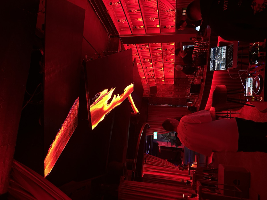 The image depicts the interior of a nightclub or lounge, identified as The Face, located in Varna, Bulgaria. The scene is illuminated predominantly by a pervasive red light, creating a distinct ambiance.
In the foreground and mid-ground, the space is occupied by several individuals. To the immediate right, partially visible from the rear, a person wears a dark hoodie with white text on the back, operating a professional DJ setup comprising a laptop computer and two controller units positioned on a low table. In the mid-ground, slightly left of center, another individual, likely male, wearing a light-colored, short-sleeved top and dark trousers, stands with their back to the viewer, facing towards a curved bar counter.
The bar counter, stretching across the right side of the image, is equipped with multiple bar stools. Behind the counter, a wall features several tiers of illuminated shelving, displaying numerous bottles. At least three other individuals, presumed to be bartenders or staff, are observed behind the bar, engaged in various tasks, their movements and identities indistinct due to the lighting.
The ceiling is dark, from which two irregularly shaped, horizontal panels are suspended. These panels display dynamic, abstract patterns of bright orange and yellow, resembling flowing lava or fire. To the left side of the room, floor-to-ceiling red drapes are visible, along with small, circular tables featuring slender bases and illuminated, candle-like objects. A dimly lit archway or entrance, exhibiting a faint blue-green glow, is present further into the background on the left.
