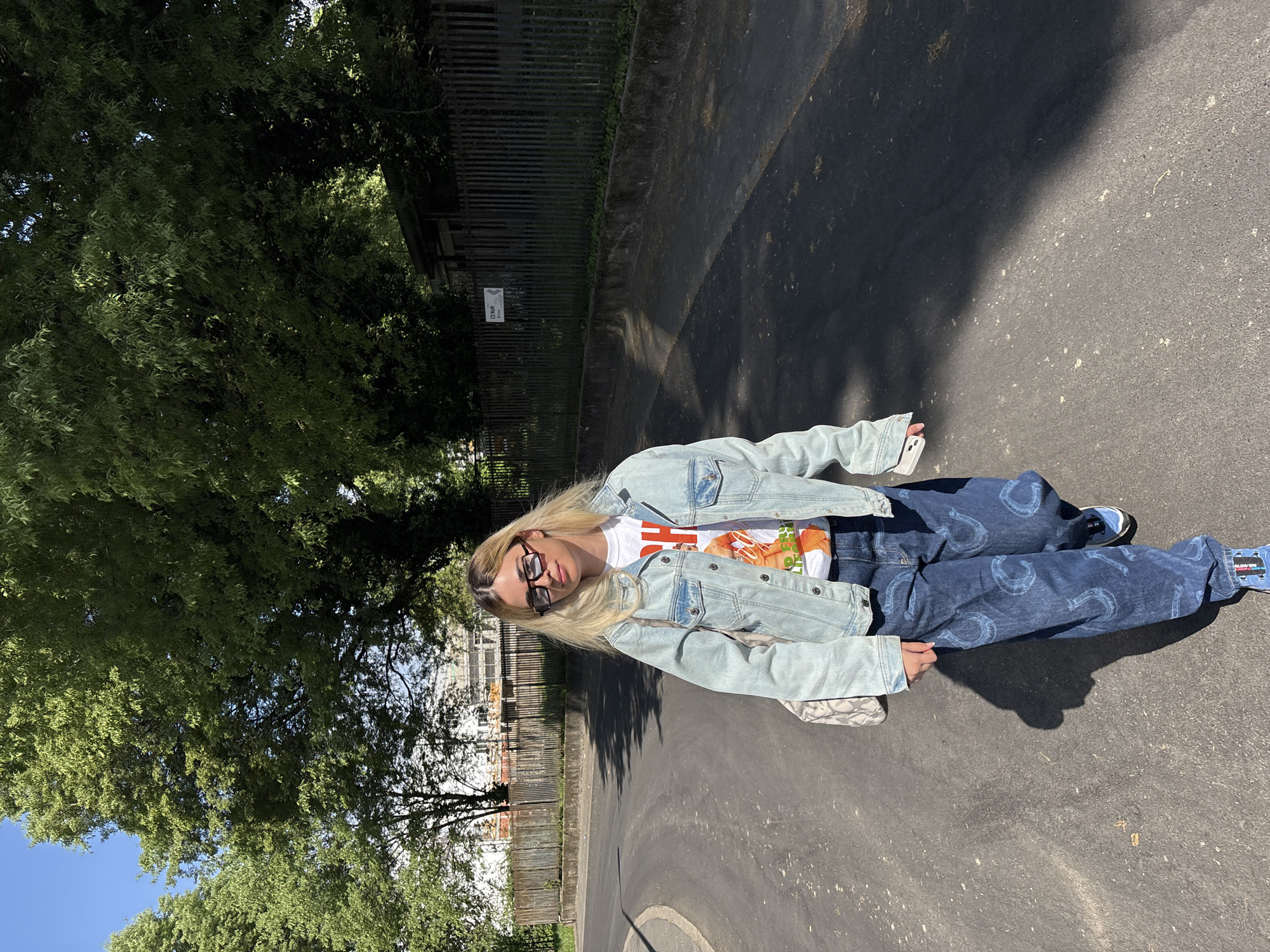 A person with long, blonde hair is the focal point of the image, wearing sunglasses, a white t-shirt with an orange graphic, a light-blue denim jacket, and dark-blue jeans with a horseshoe pattern. The person is standing on a paved surface, likely a sidewalk or road. To the left of the person is a large tree with dense green foliage. In the background, buildings and a fence can be observed. The lighting indicates it is daytime. The image appears to be rotated. There are no identifying features to confirm that this picture was taken in Milan, Italy.