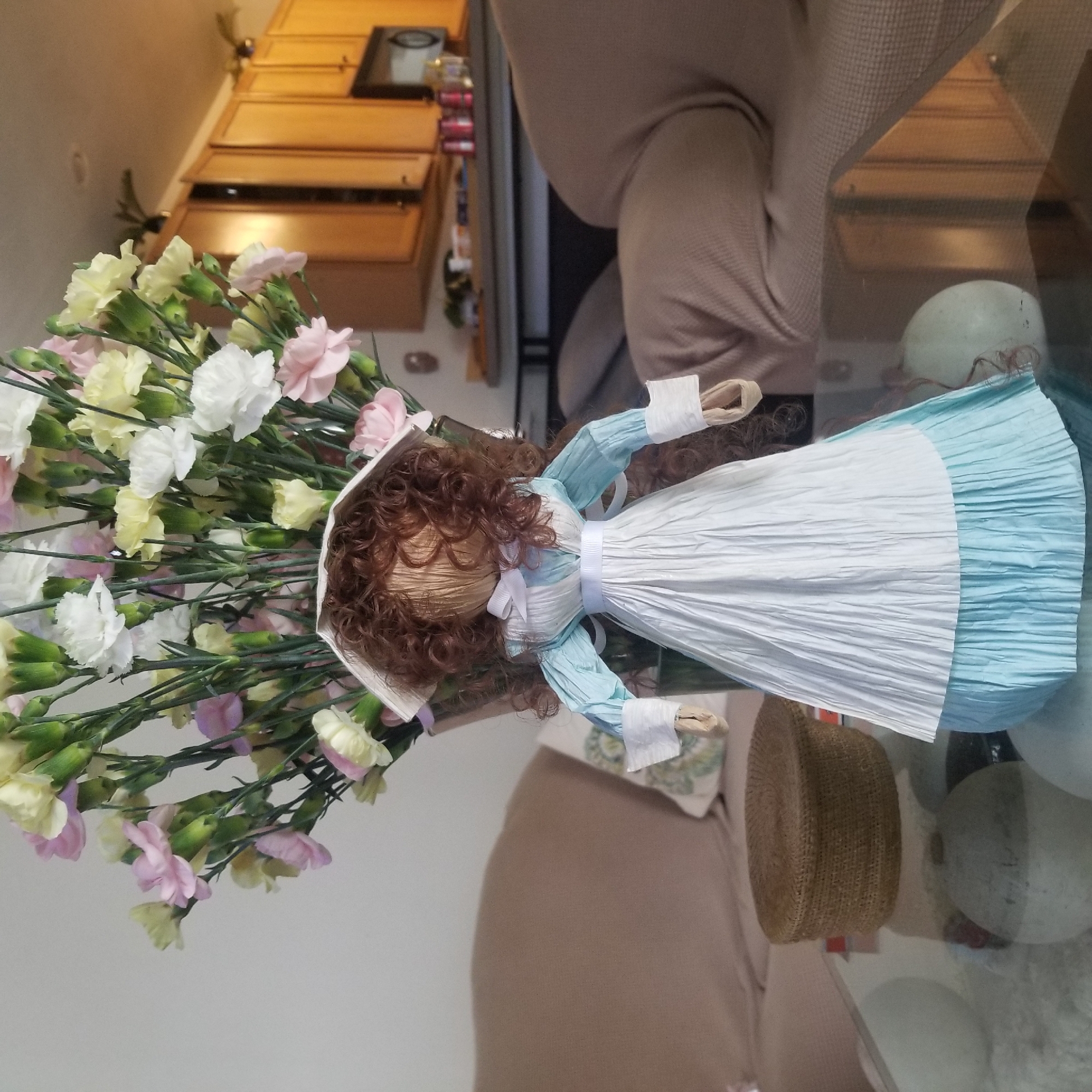 The image depicts a craft doll dressed as a nurse, standing on a glass coffee table. The doll has curly brown hair, a white cap, and a blue and white dress. Behind the doll, a large bouquet of pink, white, and yellow carnations is in a clear vase. The coffee table also holds a small woven basket and several round, light-colored decorative objects. In the background, a beige sofa is visible, and further back, a kitchen area with light wooden cabinets, a microwave, and a countertop with various items. The scene appears to be set in a home living room or dining area. There is no discernible indication of the city of Virginia Beach, United States of America.