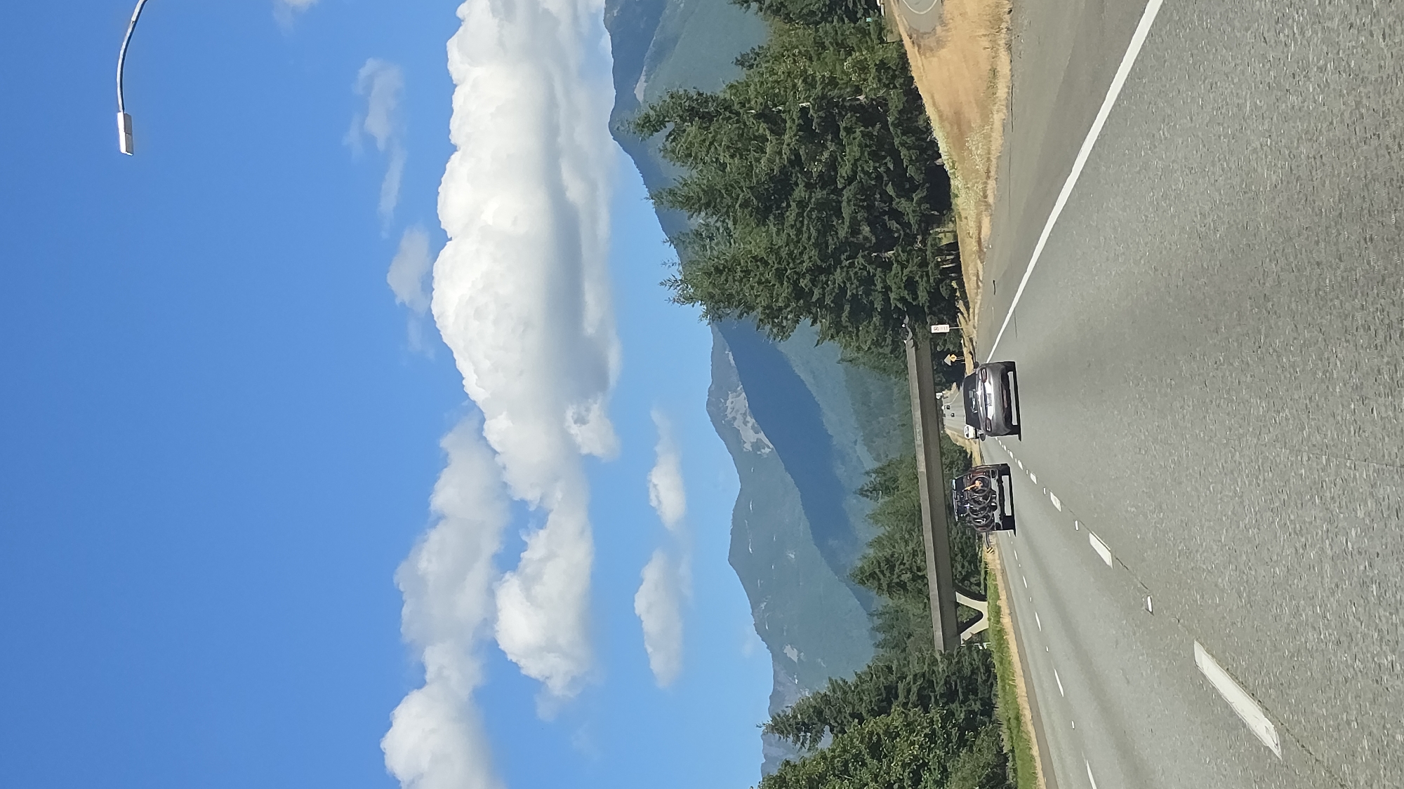 The image depicts a view from a vehicle traveling on a multi-lane highway. In the foreground, the asphalt road is marked with white lines. Ahead, two vehicles are visible: a dark-colored SUV with bicycles mounted on the back, and a silver sedan. A concrete overpass stretches across the highway in the middle ground. Beyond the overpass, a dense forest of evergreen trees lines the right side of the road. In the background, a range of majestic, tree-covered mountains stretches across the horizon, with some rocky, bare patches visible on their slopes. The sky is a clear, vibrant blue, dotted with fluffy white cumulus clouds. A modern street lamp is visible in the upper right corner of the frame, extending from a curved pole. This scene is consistent with the landscape found in the vicinity of North Bend, Washington, known for its mountainous terrain and proximity to highways.