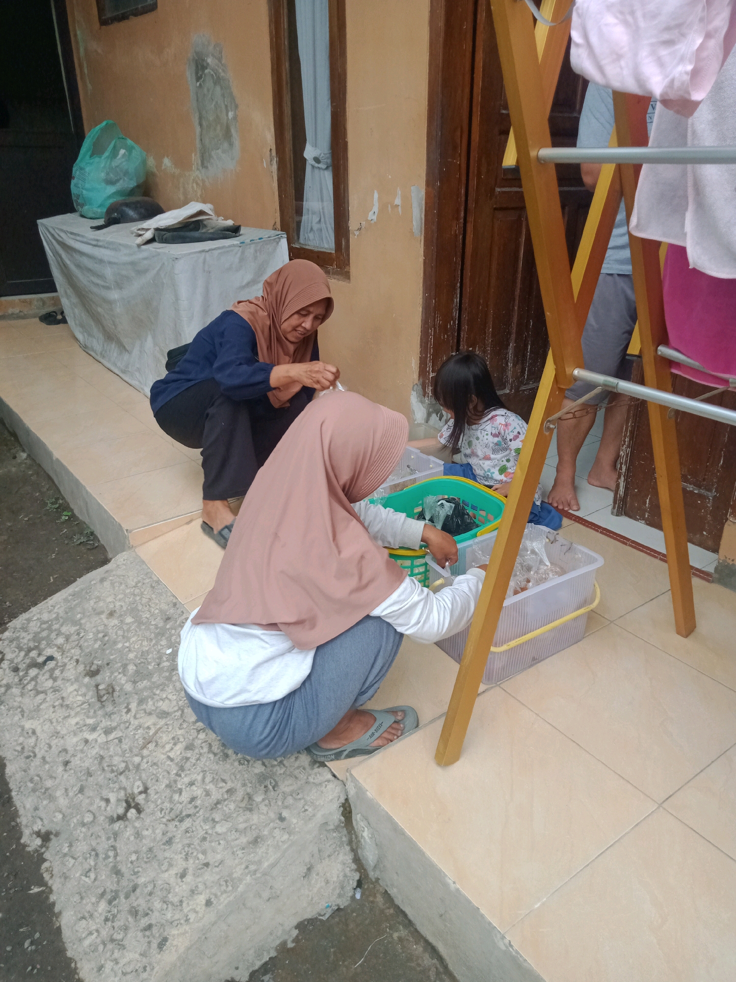 In Cintaraja, Indonesia, two women and a young girl are gathered outdoors on a tiled patio near a building. One woman, wearing a blue long-sleeved shirt and black pants, crouches on the left. She is looking down and appears to be holding a plastic bag. In front of her, another woman, wearing a beige hijab, white shirt, and grey pants, is also crouched, facing away from the camera. She is interacting with a green plastic basket filled with items. To the right of the basket, a young girl with long black hair, wearing a white and pink patterned shirt, sits on the ground. She is also looking at the basket. Adjacent to the basket is a clear plastic container holding more items, some of which are in plastic bags. A wooden drying rack with clothes hanging on it stands to the right of the scene. The background shows a light brown wall with some discoloration and a wooden door with white curtains visible through a window. A white-covered table with a green plastic bag and other objects is situated to the left. The ground consists of concrete and light-colored tiles.