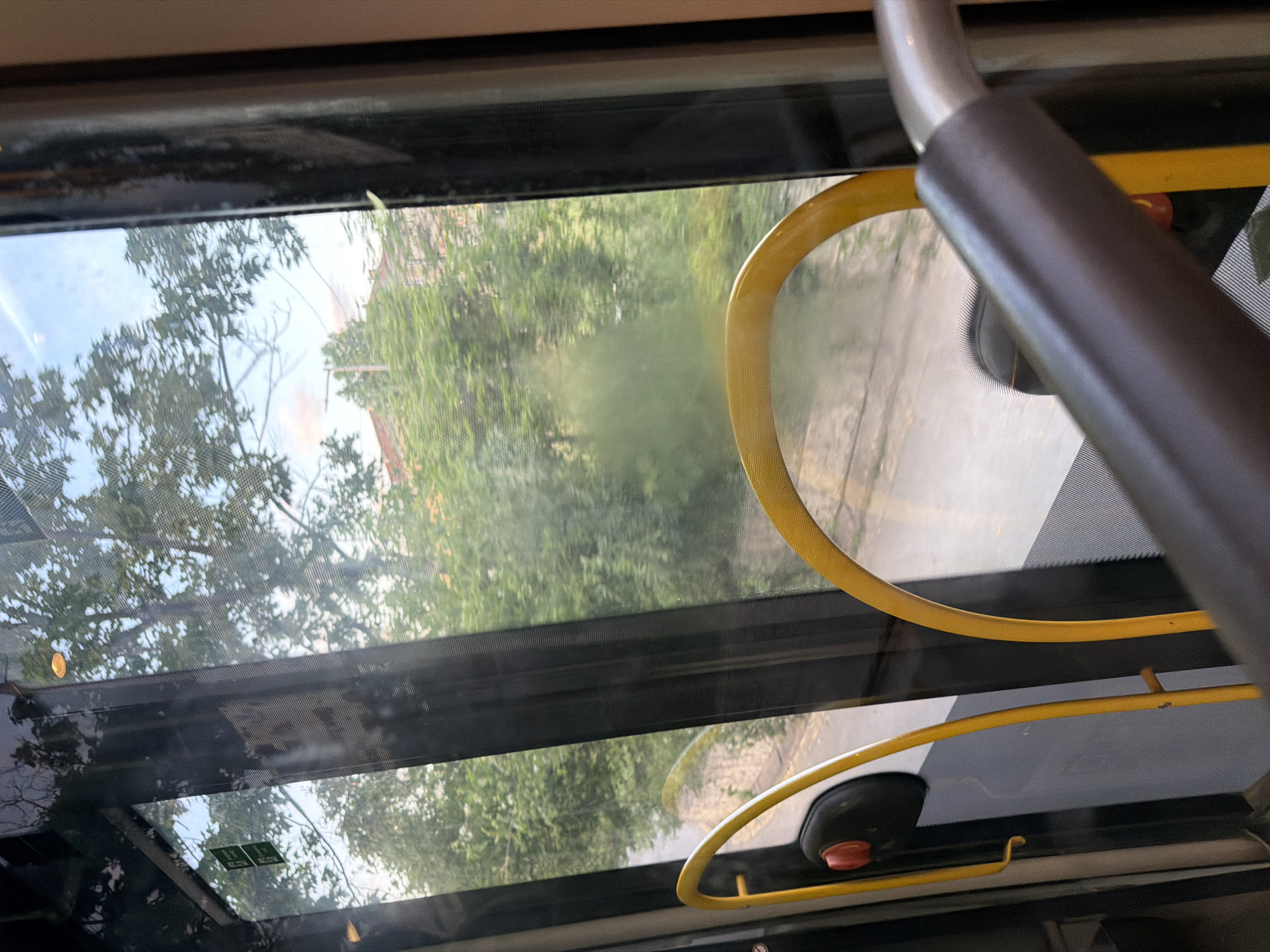 The image is taken from inside a bus in Sofia, Bulgaria. Through the bus windows, a leafy green environment with trees and buildings is visible, under a partly cloudy sky. The interior of the bus shows yellow grab bars and a grey handrail. There are no people visible in the frame. The scene suggests a daytime journey through an urban or suburban area with ample greenery.