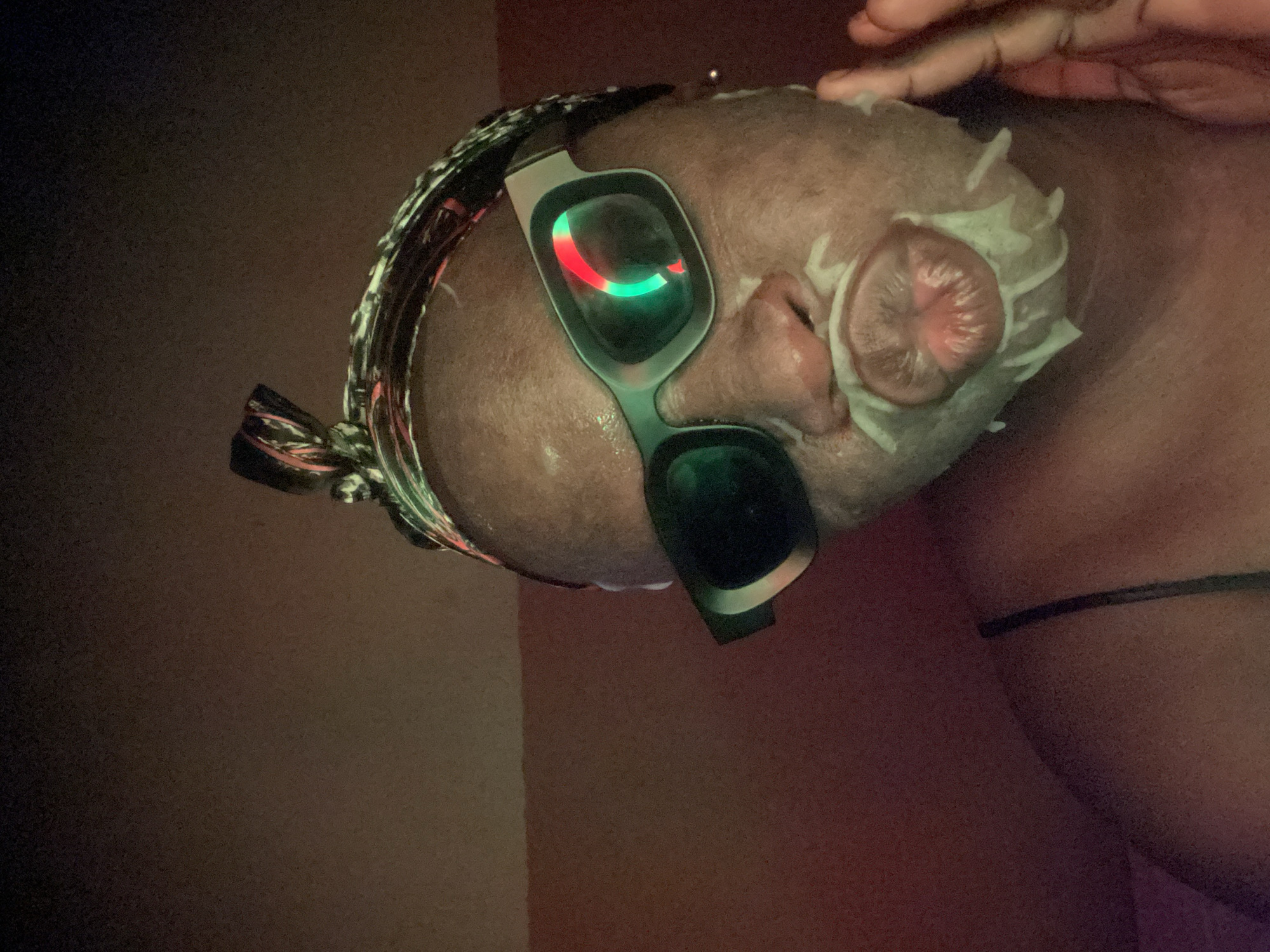 A close-up, side-angle shot shows a person's head undergoing a beauty or skin treatment in a dimly lit indoor environment in Maigizo, Nigeria. The individual has dark skin and appears to have braided hair or a protective hairstyle, partially visible along the hairline and top of the head. They are wearing specialized goggles with reflective lenses that prominently display glowing red and green lights, indicating a possible light therapy session.
A white, creamy or foamy substance, likely a facial mask or topical treatment, is applied to the side of the face, covering the skin around the ear and extending onto other facial areas. A hand, possibly belonging to the person or an aesthetician, is visible in the upper right corner of the frame, seemingly applying or adjusting the treatment. The background is dark and indistinct, featuring subtle reddish-brown hues, reinforcing the intimate and focused nature of the treatment setting. No text is visible in the image.