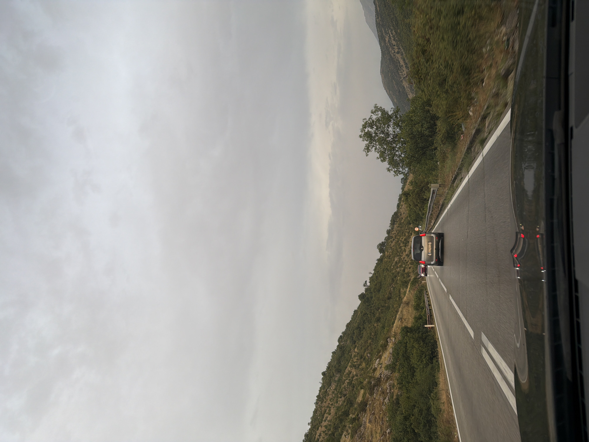 The image depicts a two-lane road in the city of Cerovo, Montenegro, on an overcast day. The perspective is from inside a vehicle, looking forward. The road surface appears wet, indicating recent rain. Two cars are visible on the road ahead: a silver SUV in the right lane and a red car further ahead in the left lane. The road is bordered by a steep, green, and rocky hillside to the left and a dense line of trees and bushes to the right. The sky is uniformly gray and cloudy. The foreground shows the hood and windshield of the vehicle from which the photo is taken.