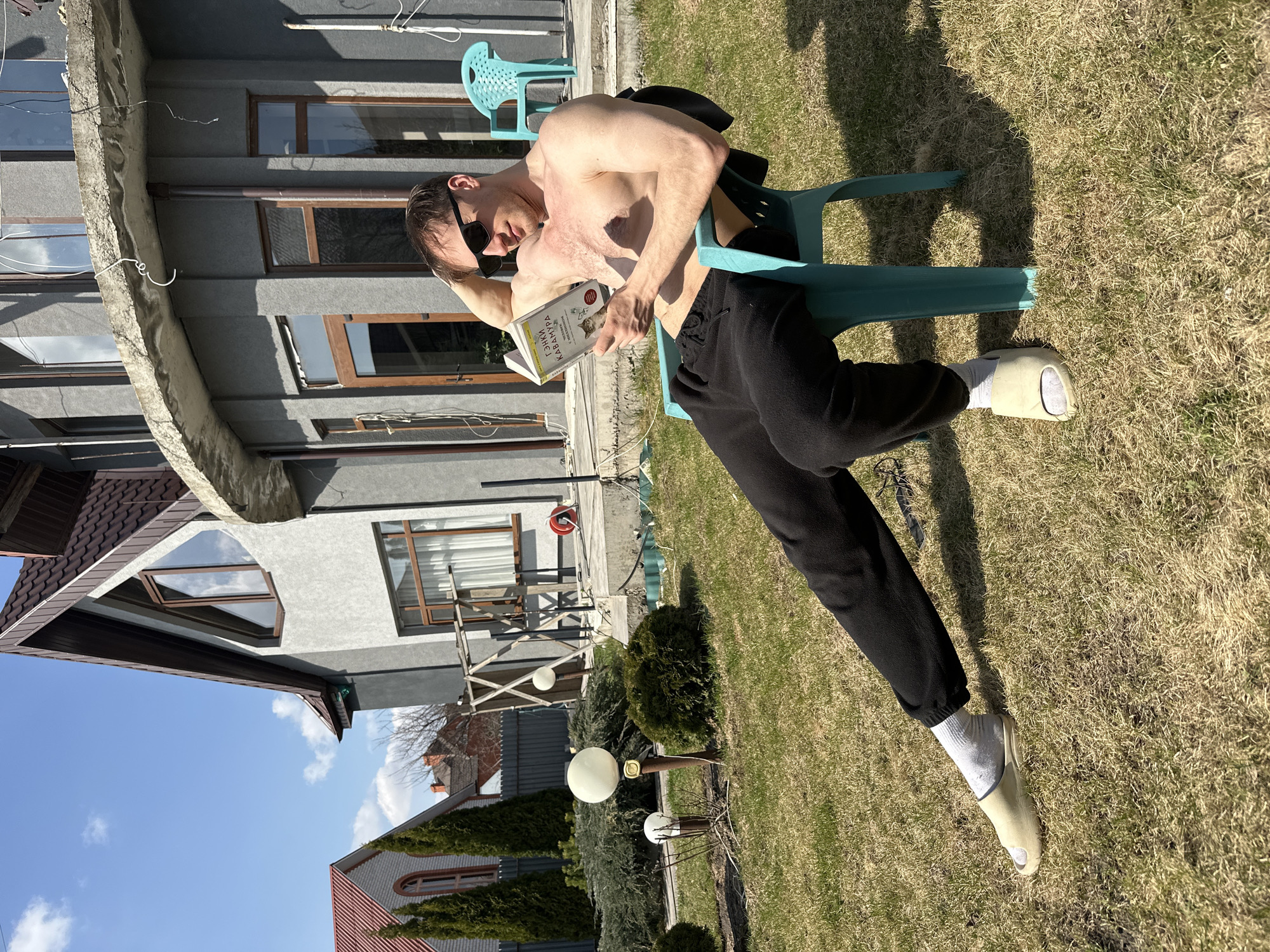 A shirtless man wearing sunglasses and black sweatpants, sits on a blue plastic chair outdoors on a sunny day, and is reading a book. He is also wearing white socks and tan-colored slides. He is positioned on a lawn with grass that is mostly brown and some green. In the background is a gray house with brown trim and a reddish-brown roof. The scene appears to be in a residential area. There is no verifiable information in the image that confirms this is the city of Kursk, Russia.