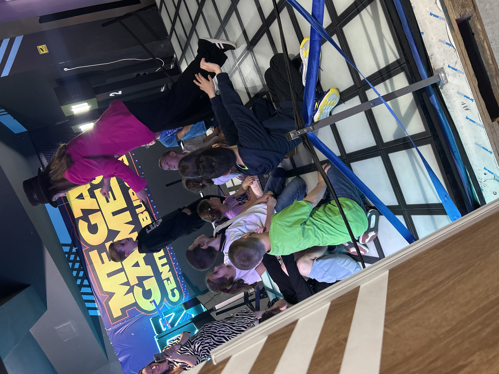 A large group of children are inside what appears to be an upside-down room. The walls, ceiling, and floor are decorated as if they were once in a regular upright orientation. A sign reading "Mega Games Center" is seen. The location appears to be indoors, possibly an entertainment center in Burgas, Bulgaria, given context clues from the images' names. Some children are wearing shoes that suggest their feet are on the ceiling. They are interacting with the features as if defying gravity. Blue straps and metal fixtures are attached to the ceiling.