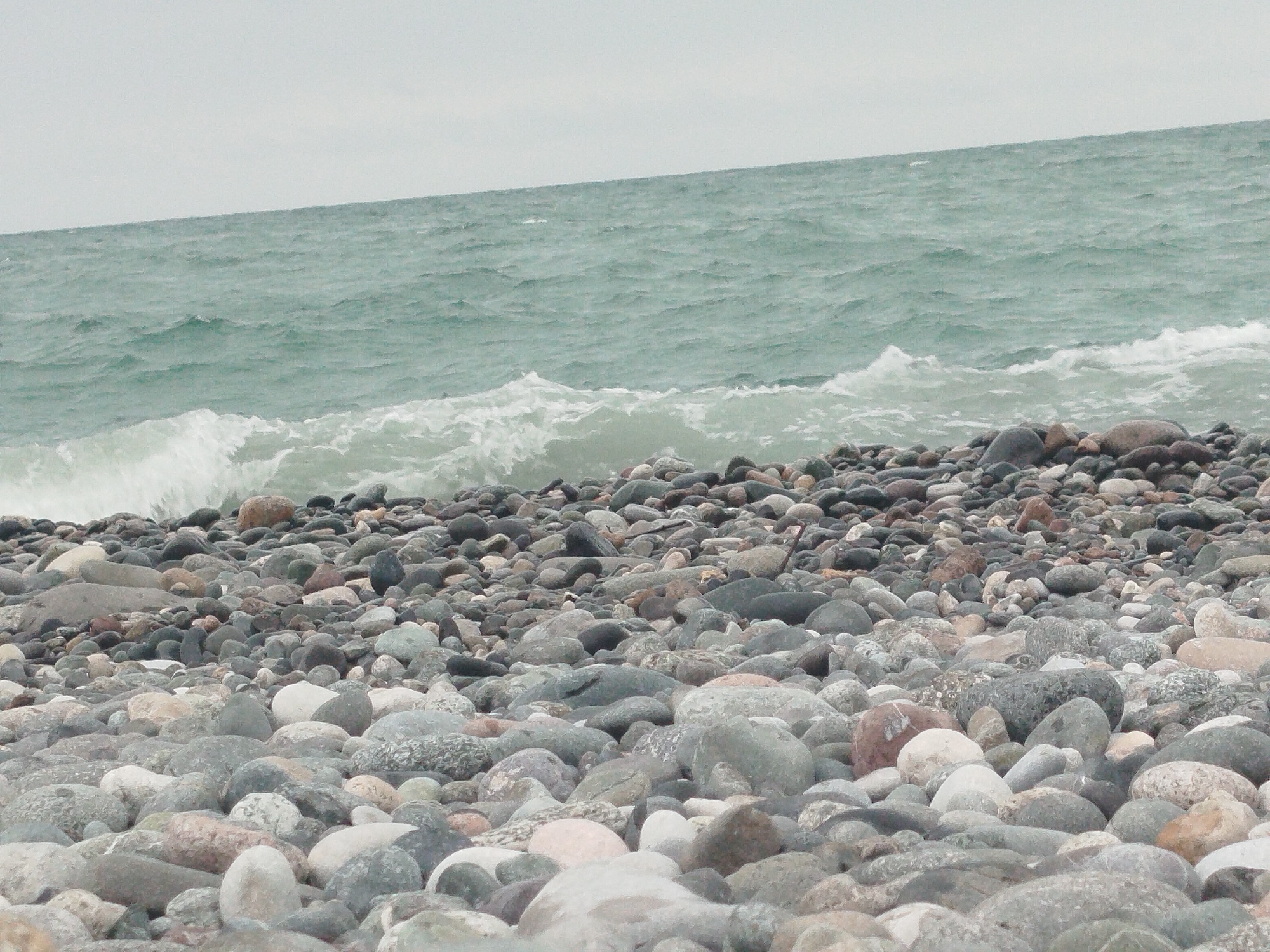 The image depicts a rocky beach with the ocean in the background. The foreground is filled with a dense arrangement of various sized and colored stones. The sea is a muted green with visible waves breaking and receding. The sky is overcast and a uniform light gray, suggesting a cloudy day. There are no people or man-made structures visible in the image. The scene appears to be a natural coastal landscape.