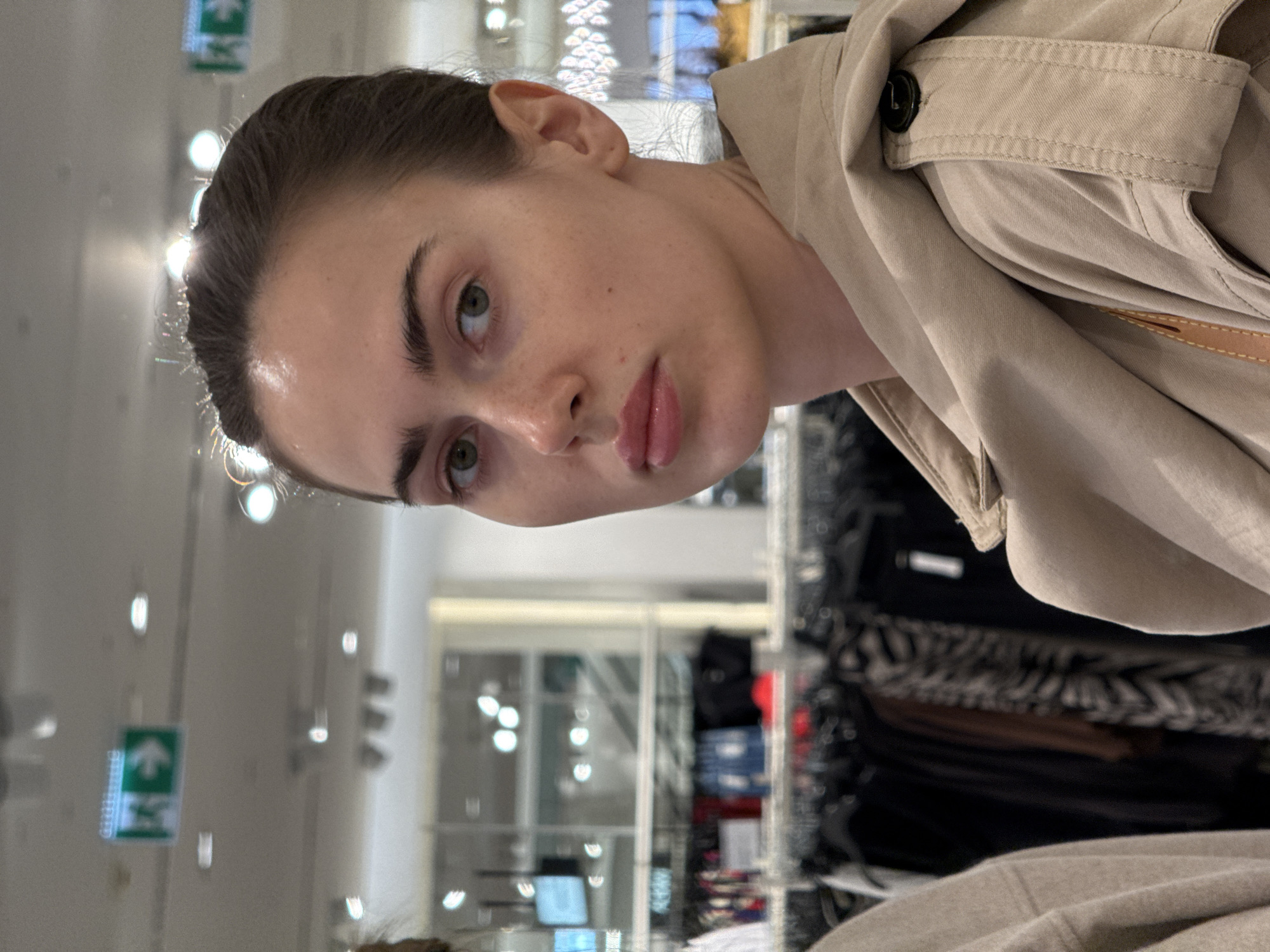 A woman with fair skin, dark hair pulled back, and green eyes is positioned in a retail environment. She is wearing a tan trench coat with a button visible on the collar. The store's interior is visible in the background, featuring clothing racks filled with dark-colored garments. Above and behind the woman are lights and signs, including a green emergency exit sign with a white arrow pointing upwards and to the left. The location appears to be indoors in the city of Unknown, Unknown.
