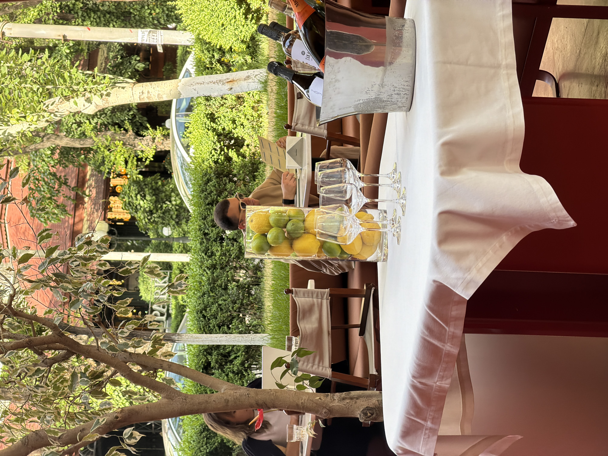 The image shows an outdoor dining area, likely a restaurant patio. A table draped with a white tablecloth dominates the foreground. On the table are a display of lemons and limes in a clear rectangular container, several wine glasses, and a champagne bucket with bottles of wine. A man wearing sunglasses is seated at the table, looking at a menu. In the background, there is lush green foliage and trees. A tree with variegated leaves frames the left side of the scene. A red building is partially visible in the upper left background. Another person is seated at a table near the foreground tree. Based on the street lamp with Armenian text on the left, this image is likely from a restaurant in Yerevan, Armenia.