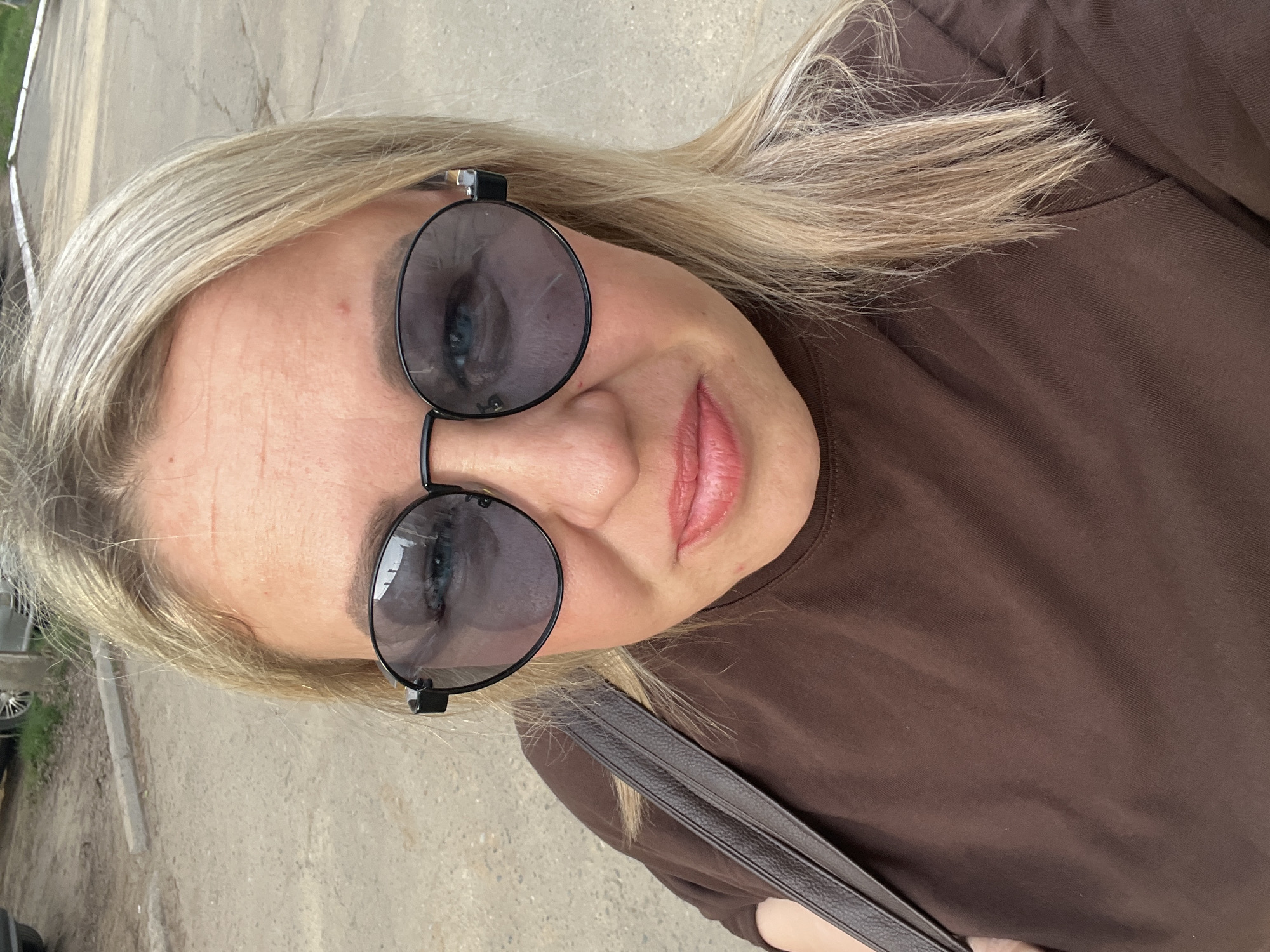 A selfie, rotated counter-clockwise, depicts a blonde woman wearing round, dark sunglasses and a brown t-shirt. The reflection in her sunglasses shows an outdoor environment. A dark leather strap, possibly from a bag, is visible across her chest. The background shows a concrete or paved surface. The location cannot be determined with the given information.