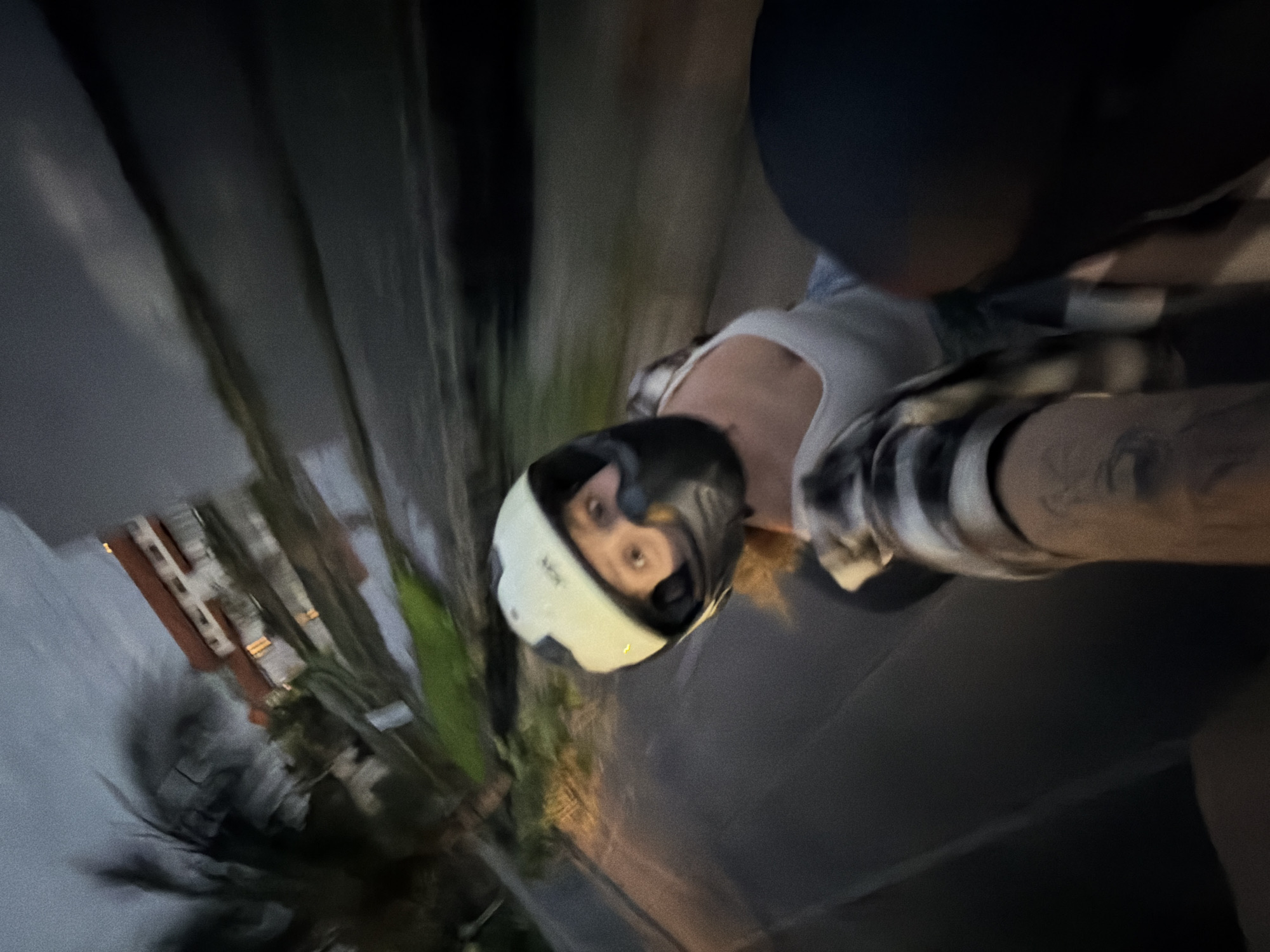 A blurry, tilted photograph captures a person wearing a white helmet with a dark visor, possibly riding a moped or motorcycle at night. The person is wearing a light-colored tank top and a plaid shirt. The person has visible tattoos on their arm. The environment appears to be an urban street in North Kuta, Indonesia, with blurred buildings, trees, and street lights suggesting motion.