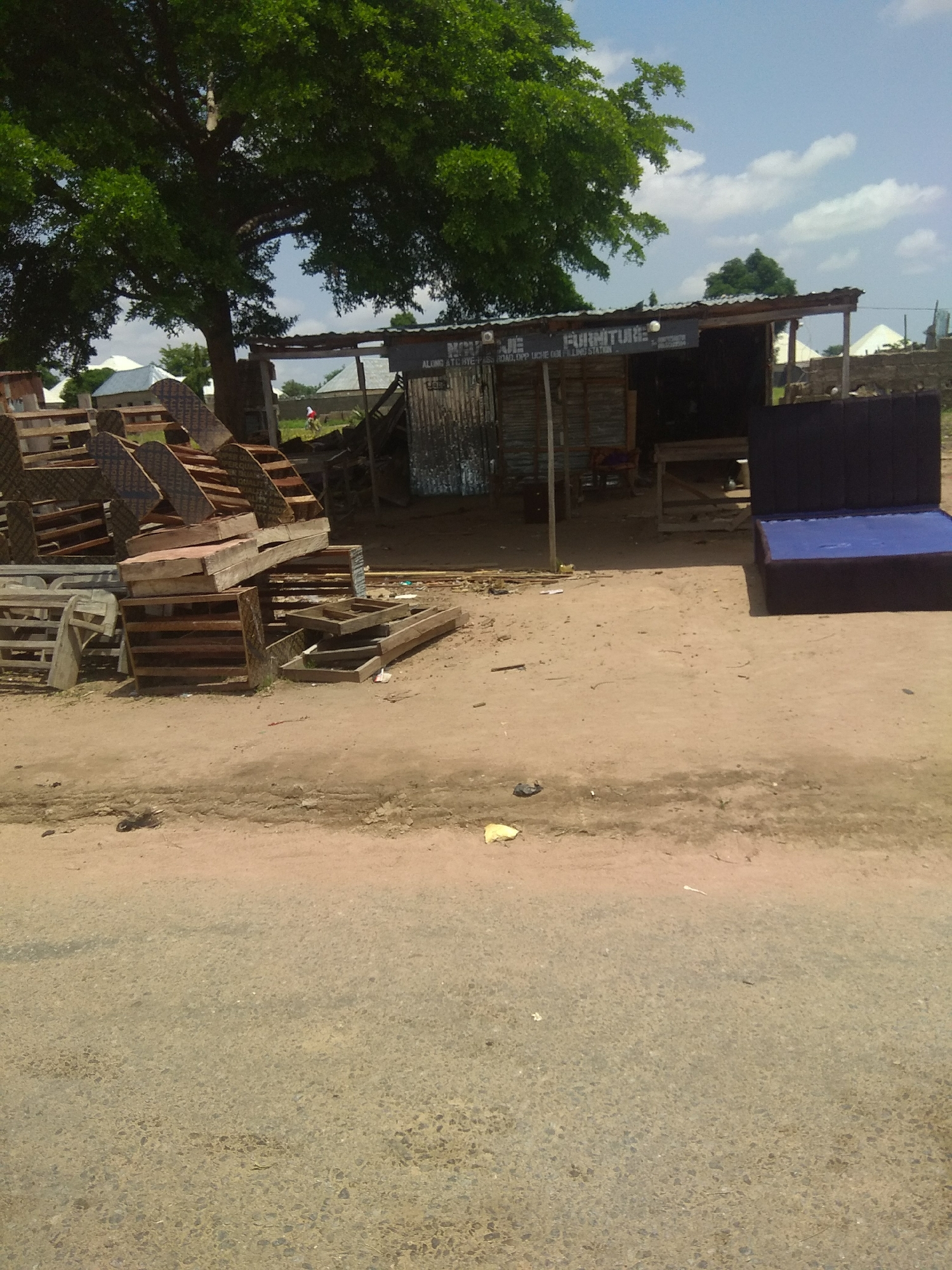 This image shows a street-side furniture shop in Jalingo, Nigeria. The shop itself is a simple structure with a corrugated metal roof and walls, open on one side to display its wares. In front of the shop, there is a large pile of wooden pallets and construction materials. A dark blue upholstered bed frame with a blue mattress is displayed prominently to the right of the shop.
The scene appears to be outdoors during the day, with a bright sky visible through the leaves of a large tree that overhangs the area. The ground is dusty and appears to be unpaved, suggesting a rural or semi-urban setting. There are a few pieces of debris scattered on the ground, including a yellow plastic bag and some small black objects.
A sign is visible above the shop entrance with text that reads "FURNITURE". Partially visible text below it includes "ALIMI" and "ROAD, OPP UCHE IY... FILLING STATION". There is a person in the background, wearing a red shirt and white pants, walking away from the shop. The overall impression is one of a functional, no-frills business operating in a warm climate. The weather appears to be clear and sunny.