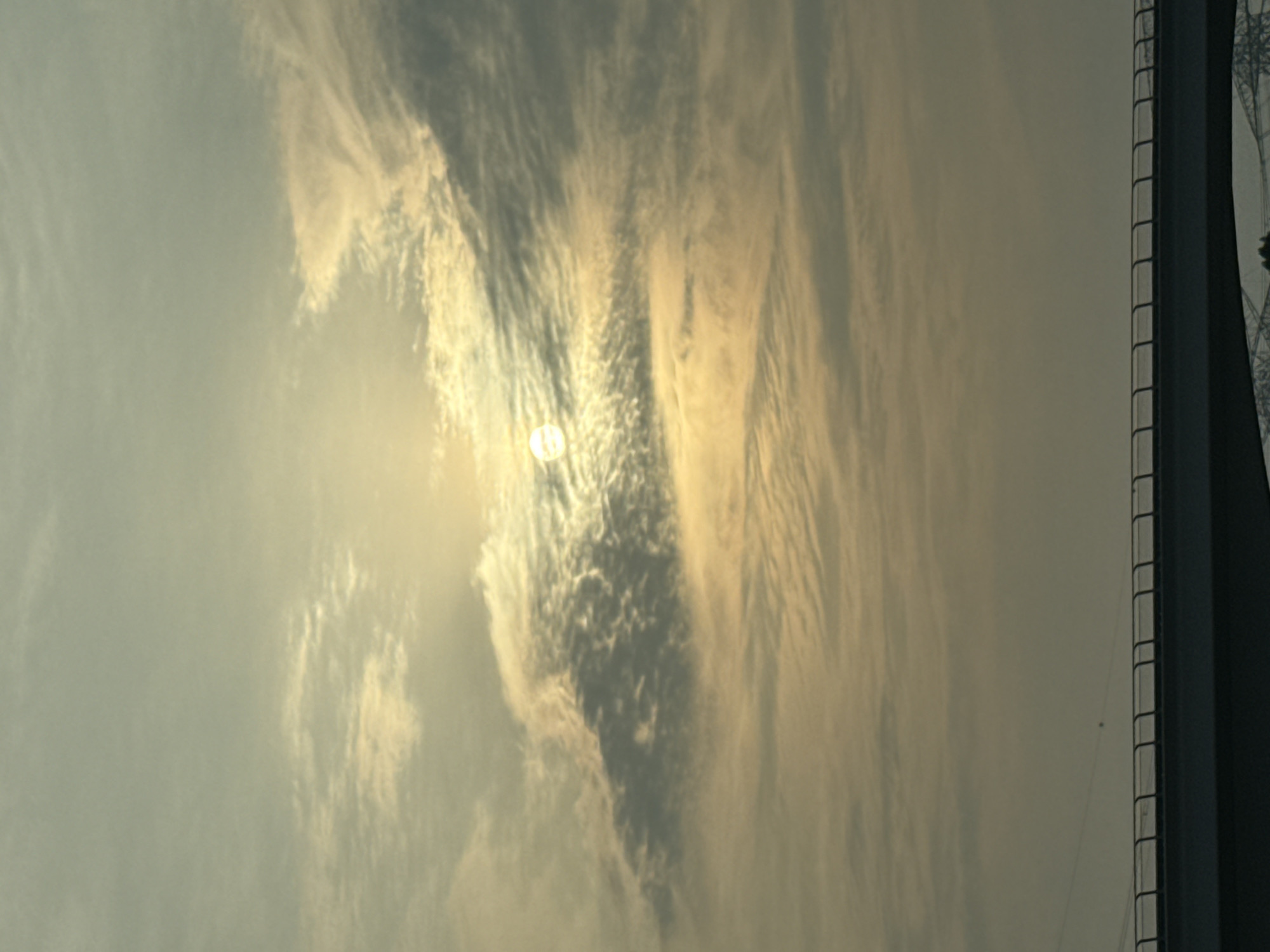 The image depicts a sky filled with diffused clouds, with the sun visible as a bright, circular orb partially obscured by them. The sun appears to be low in the sky, suggesting either sunrise or sunset. In the foreground, a portion of a bridge is visible, characterized by a dark, solid barrier with a railing on top. The overall lighting and atmospheric conditions suggest a hazy or overcast day, common in many parts of the world, including Dubai, United Arab Emirates. However, no specific landmarks or distinct features are present in the image that would allow for precise identification of its location within Dubai. The scene is devoid of people, animals, or any discernible activity.