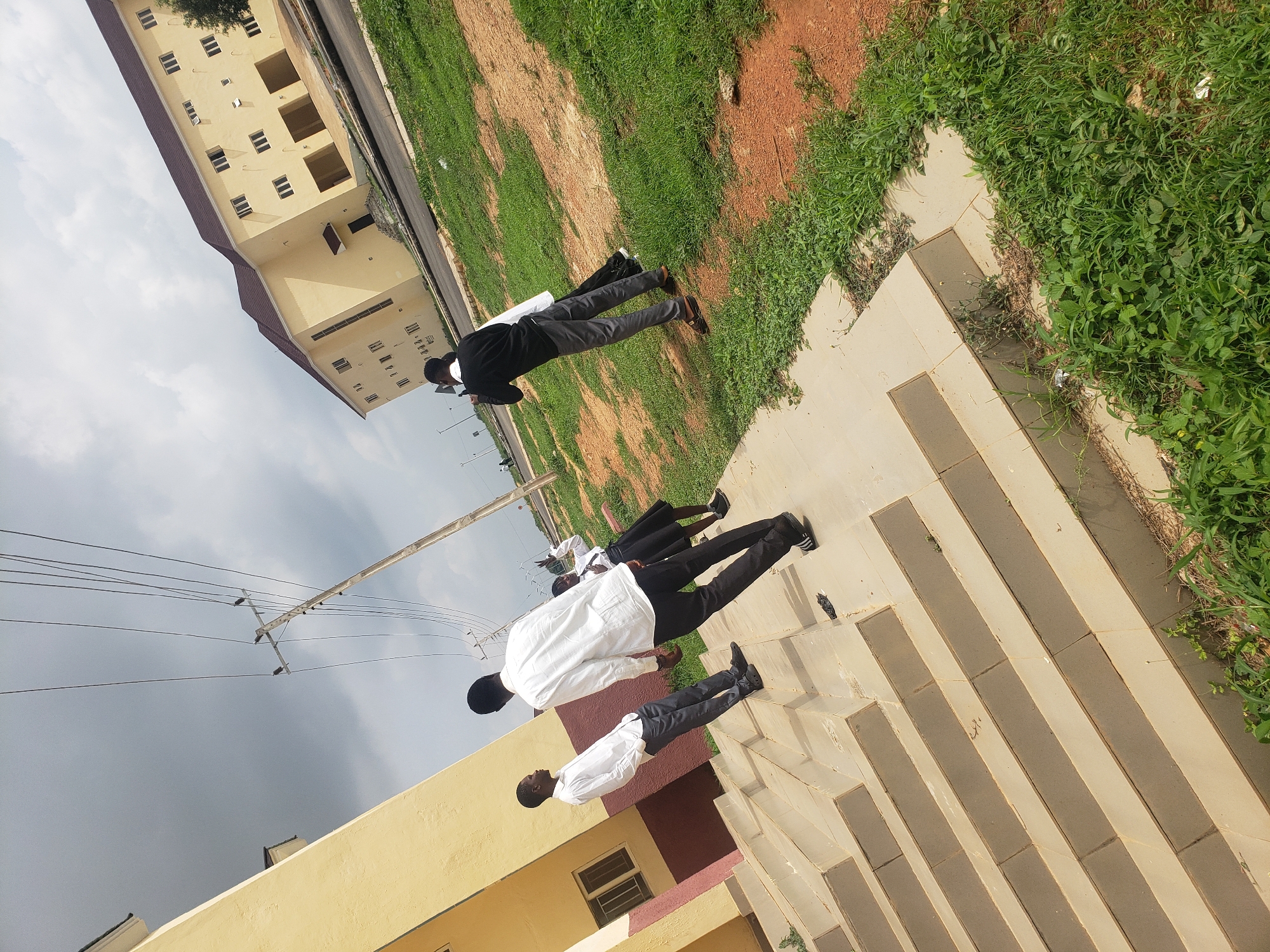 In the city of Gumel, Nigeria, on a bright but overcast day, three individuals are observed near a light yellow building with a dark roof. The building appears to be a two-story structure with numerous windows. In the foreground, a set of light-colored, wide stairs leads up to an entrance.
On the stairs, two young men are positioned. The one closer to the viewer is wearing a white long-sleeved shirt and dark trousers, facing away from the camera and looking towards his companion. The other young man, further up the stairs and to the left, is also in a white long-sleeved shirt and dark trousers. He is facing left, away from the camera, and appears to be looking in the same direction as the first man.
Behind them, further up the stairs and to the right, a young woman is visible. She is wearing a white long-sleeved shirt and a dark skirt. She is looking towards the camera, and her right hand is raised in what appears to be a peace sign gesture.
In the background, the light yellow building continues to dominate the scene. To the right of the building, a road is visible, with sparse green grass and reddish-brown dirt patches alongside it. Power lines stretch across the sky, supported by a wooden utility pole. The sky is cloudy, with patches of brighter sky suggesting a mix of sun and cloud cover. The overall impression is of a casual outdoor gathering or moment of interaction between the individuals.