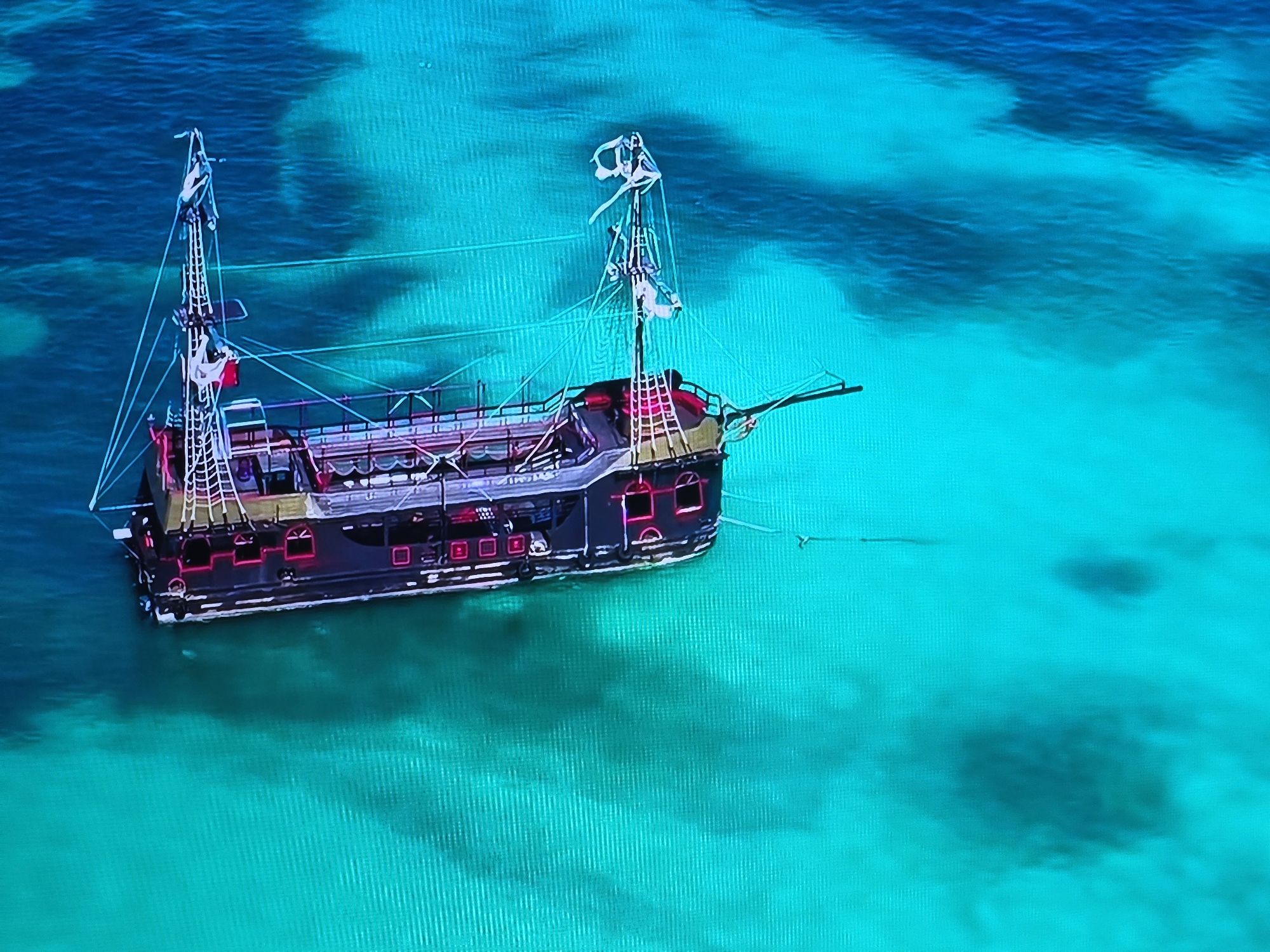 An aerial or high-angle view captures a large, dark-hulled ship resembling a pirate vessel, anchored in exceptionally clear, turquoise-colored water. The ship features a dark body, possibly black or very dark grey, accented with vibrant pink or magenta lighting around its numerous windows and along its railings. Two tall masts rise from the deck, adorned with complex rigging and white, tattered decorative elements or furled sails at their tops. One mast displays a small flag with red and white coloring.
The water surrounding the ship is translucent, revealing the sandy and variegated texture of the seafloor beneath. The color of the water ranges from a luminous light turquoise in shallower areas to a deeper blue-green where the water is slightly deeper. The scene is brightly lit, suggesting a clear day with calm conditions. No people are visible on board the ship or in the water. The image evokes a sense of tropical beauty and tranquility, likely depicting a themed tour boat or replica ship in a pristine marine environment such as a bay or lagoon. There is no visible text in the image.
