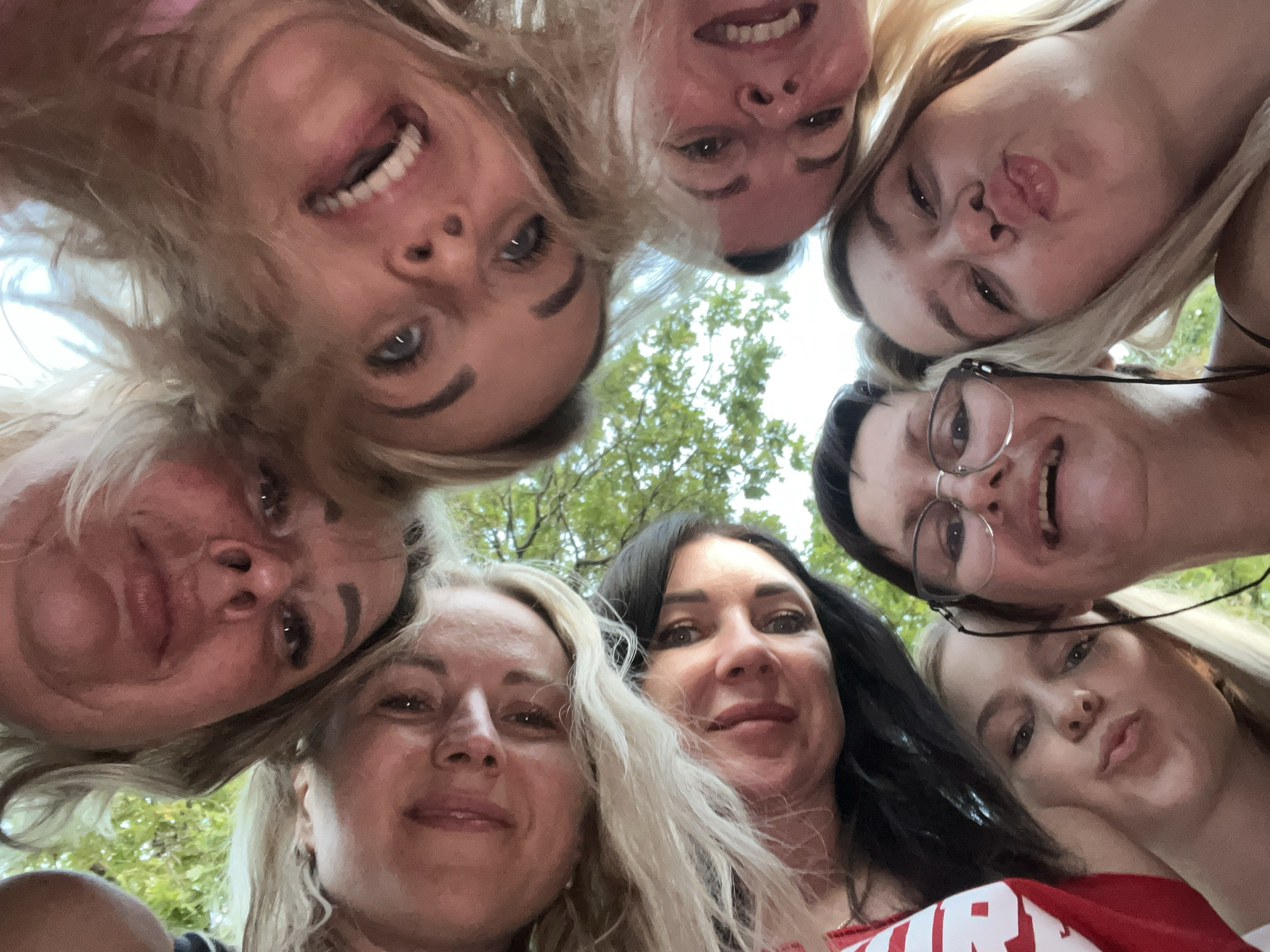 Eight people, appearing to be women and girls, are positioned in a circle facing inwards, with their faces close together and angled towards the camera in a low-angle shot. They are outdoors, as indicated by the visible green foliage and sky in the background. Some individuals have their mouths open, either smiling or making kissing gestures. Their faces and hair are prominently featured in the foreground. No specific location in the city of Shumeika, Russia, can be identified from the image.