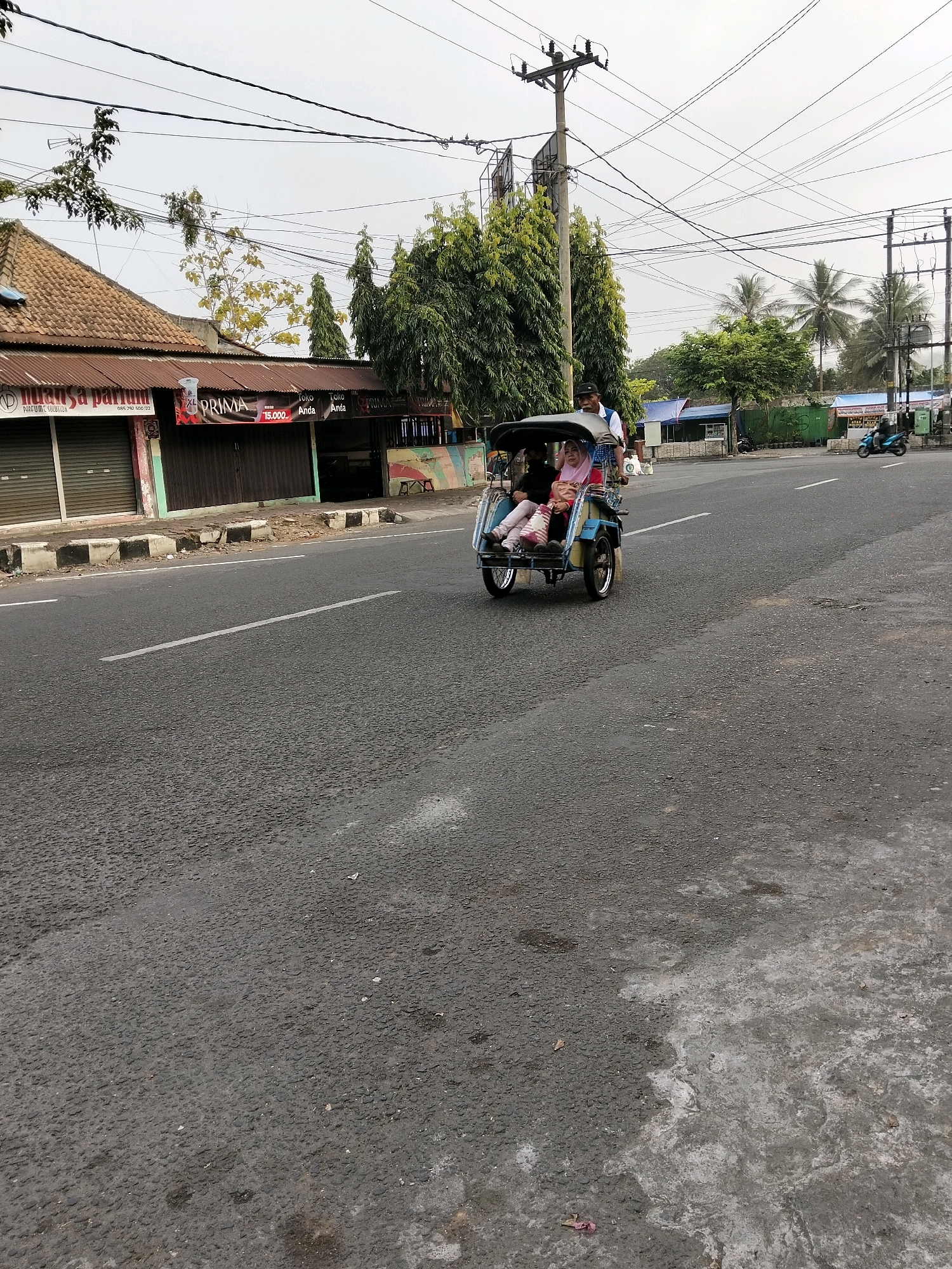 The image depicts a street scene in Purworejo, Indonesia, with a three-wheeled vehicle, often referred to as a "becak motor" or motorized pedicab, driving on the asphalt road. Two individuals are seated within the vehicle; a woman in the front, wearing a light pink headscarf and a patterned bag on her lap, and a person behind her, whose face is obscured. A man in a white shirt and dark hat is operating the vehicle. On the left side of the road, a building with a tiled roof and closed metallic shutters is visible, featuring signage including "NUANSA PARFUM" and "XL PRIMA." Further down the street, a blue motorcycle is seen in motion. The environment includes a utility pole with numerous overhead wires, palm trees, and lush green foliage. The sky is overcast, suggesting a cloudy day. The road surface shows signs of wear and tear, with some patches of discoloration.