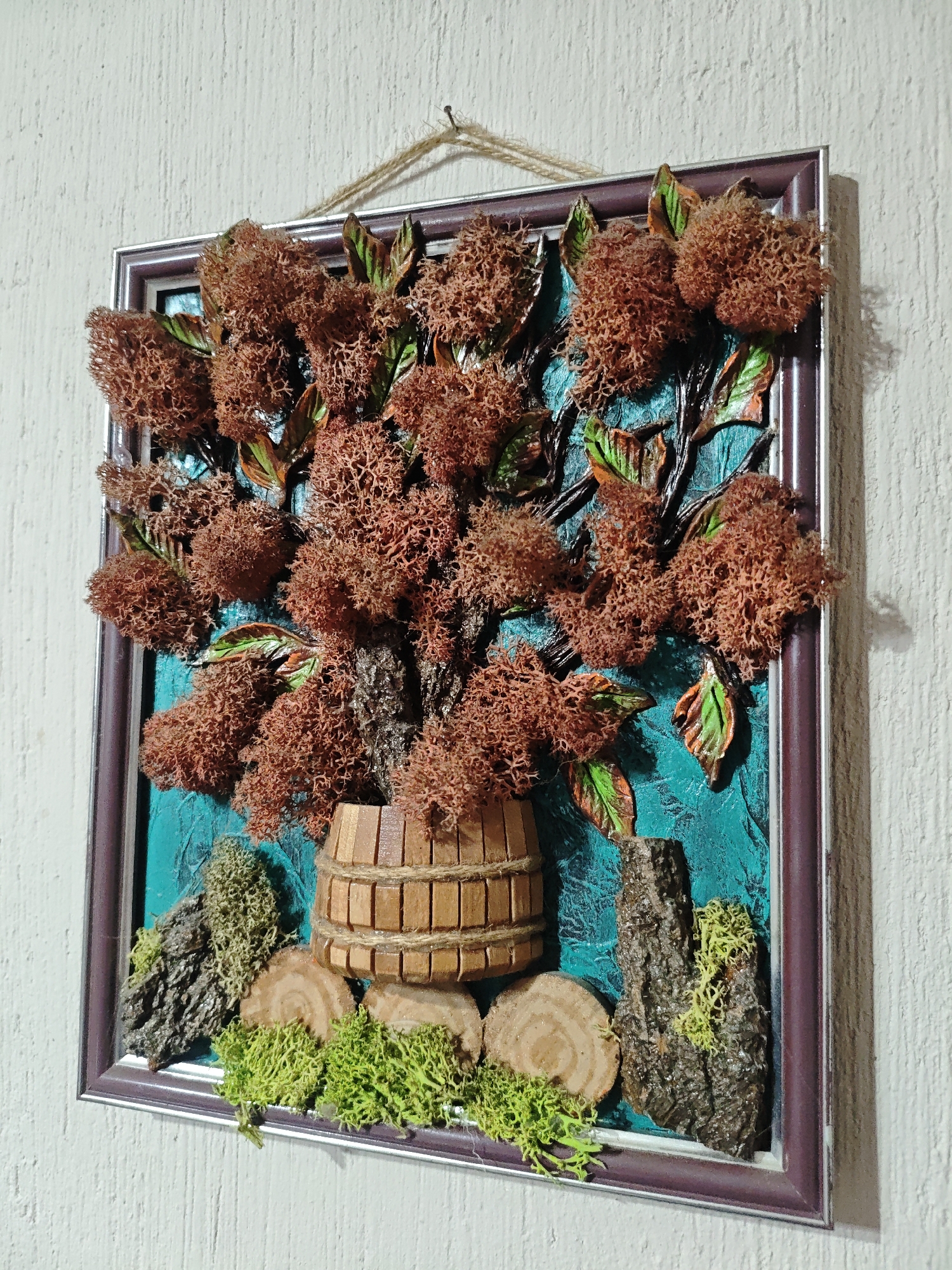 A framed artwork hangs on a white wall. The artwork features a tree with textured brown foliage and green leaves, set against a teal-colored background. The tree's trunk appears to emerge from a small wooden barrel or container. At the base of the artwork, there are pieces of wood, tree bark, and patches of green moss. The frame is a combination of silver and maroon. A piece of string is attached to the frame, suggesting it's used for hanging the artwork. The location is an interior, based on the plain wall. There is no information available on the city.