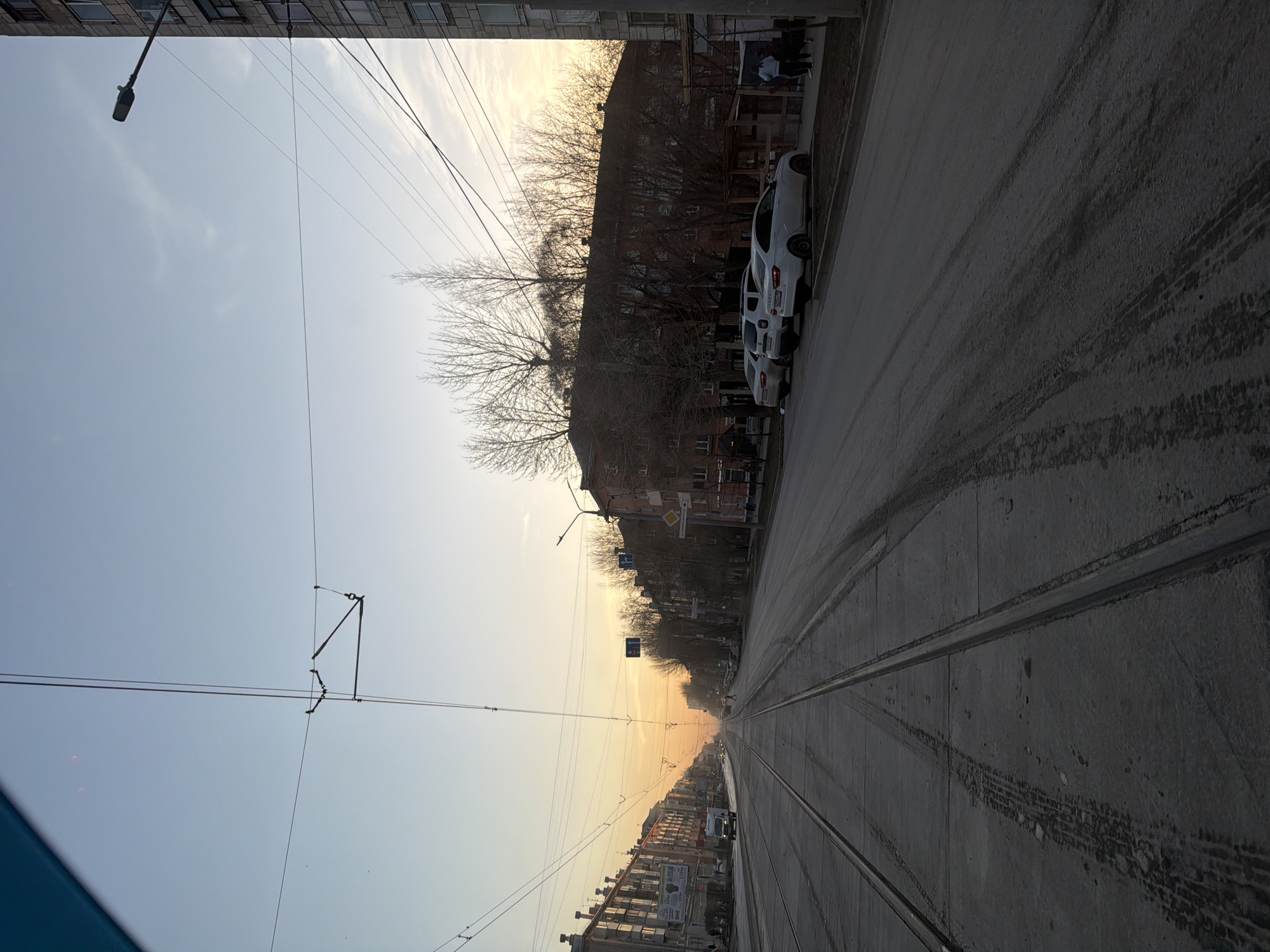 The image depicts a street view in Perm, Russia, notable for the presence of tram tracks embedded in the concrete road surface. Overhead, tram wires are visible, suspended between buildings and supporting poles. A row of white cars is parked along the roadside next to an older residential building. The sky appears bright with sunlight. There are pedestrians on the sidewalk.