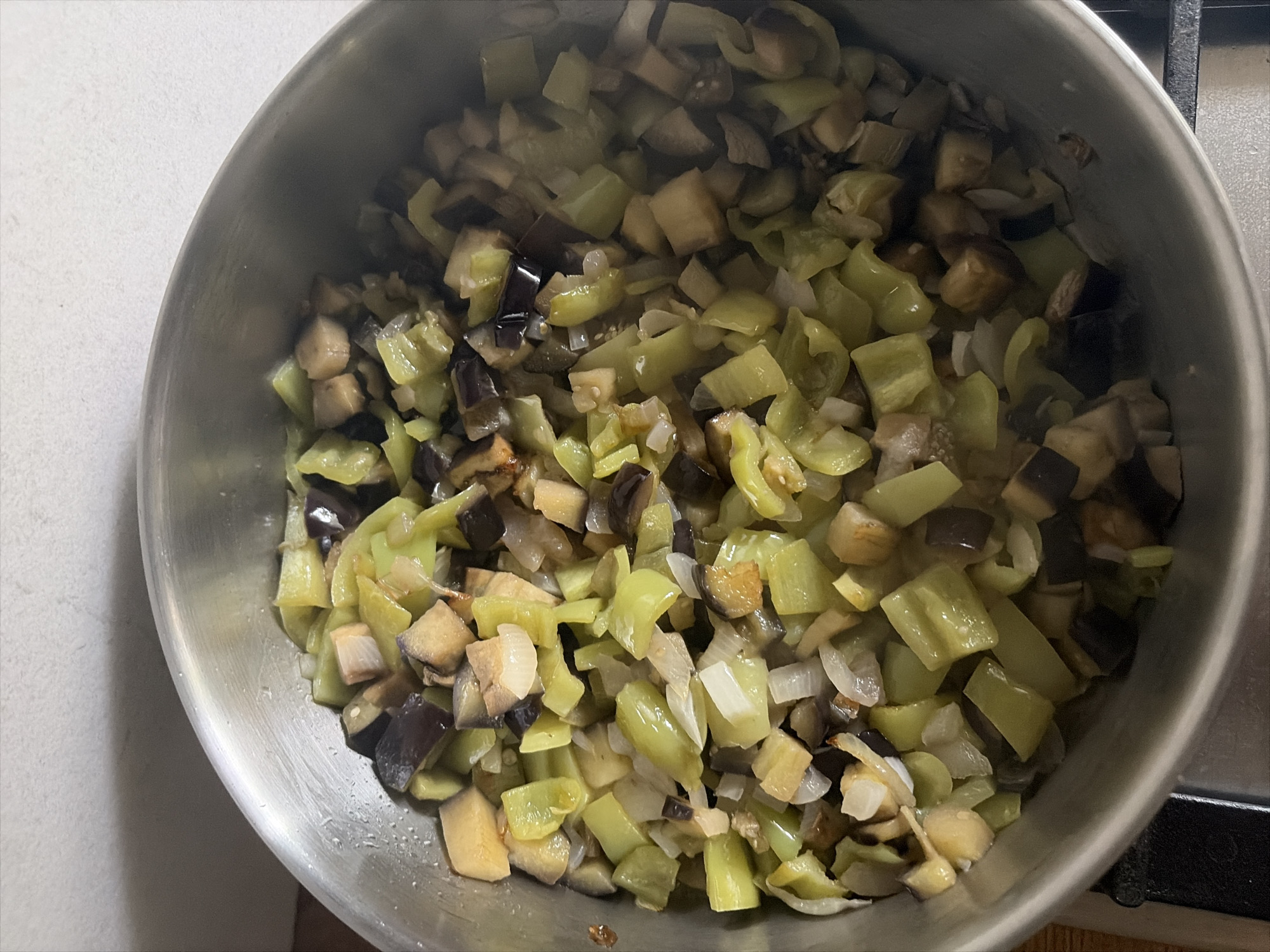 The image shows a close-up, overhead view of a stainless steel pot filled with chopped vegetables, likely eggplant and peppers, sauteing. The vegetables are cut into bite-sized cubes and appear to be lightly browned in places, suggesting they are being cooked. The pot is sitting on a black stovetop. The background is a textured, light-colored wall. There is no information about the city of Oxentea, Moldova present in the image or derivable from it.