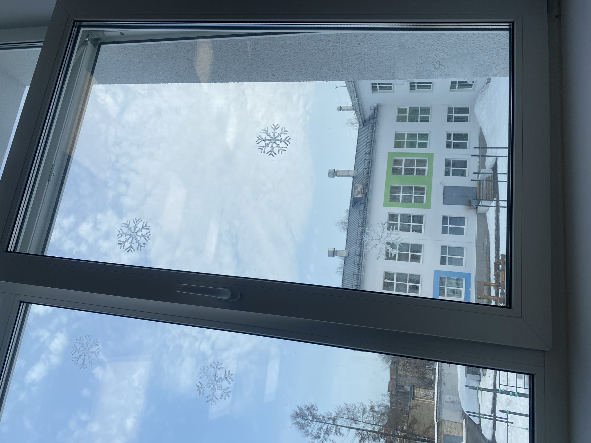 The image depicts an exterior winter scene viewed through a partially opened, white PVC-framed window, adorned with five translucent snowflake decals on its glass panes. The window's bottom frame and a white windowsill are visible at the base of the photograph.
Outside, a prominent two-story, white building with a gray pitched roof is the primary subject, located in the city of Chaikovsky, Russia. This building features multiple rectangular white windows, some of which are highlighted with colored exterior panels: two prominent green sections on the upper floor and a blue section on the lower floor. Several cylindrical, silver-colored exhaust vents or chimneys protrude from the roofline.
The ground surrounding the building is covered in snow, with a significant snow pile directly in front of the building's central section. A set of concrete steps leads to a dark gray entrance door on the ground floor, partially obscured by snow and a low metal railing. To the right of the entrance, a small wooden structure, possibly a piece of playground equipment, is visible amidst the snow. To the left of the main building, further snow-covered ground and smaller, single-story structures resembling garages or outbuildings are visible, alongside bare deciduous trees in the mid-distance. The sky above is clear blue with scattered white, fluffy clouds. No individuals are present in the scene.