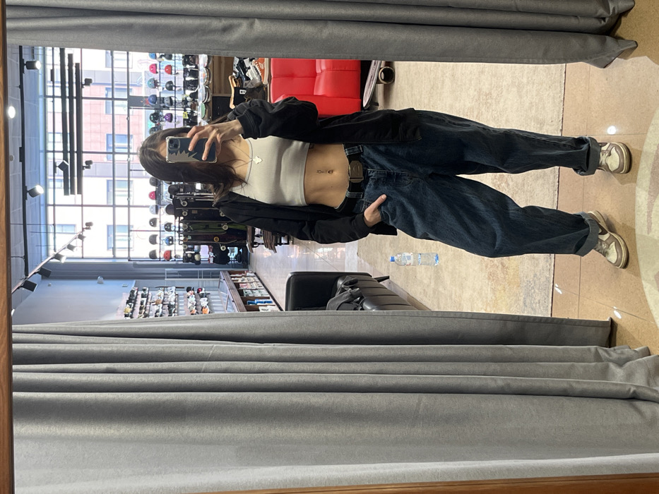 A person in a mirror selfie, standing in front of grey curtains. They are wearing a white crop top, dark jacket, and baggy blue jeans with a belt. Their phone covers part of their face. In the reflection, a store interior is visible with shelves, windows, and helmets on display. A red couch sits in the background. A water bottle lies on the floor near a suitcase.