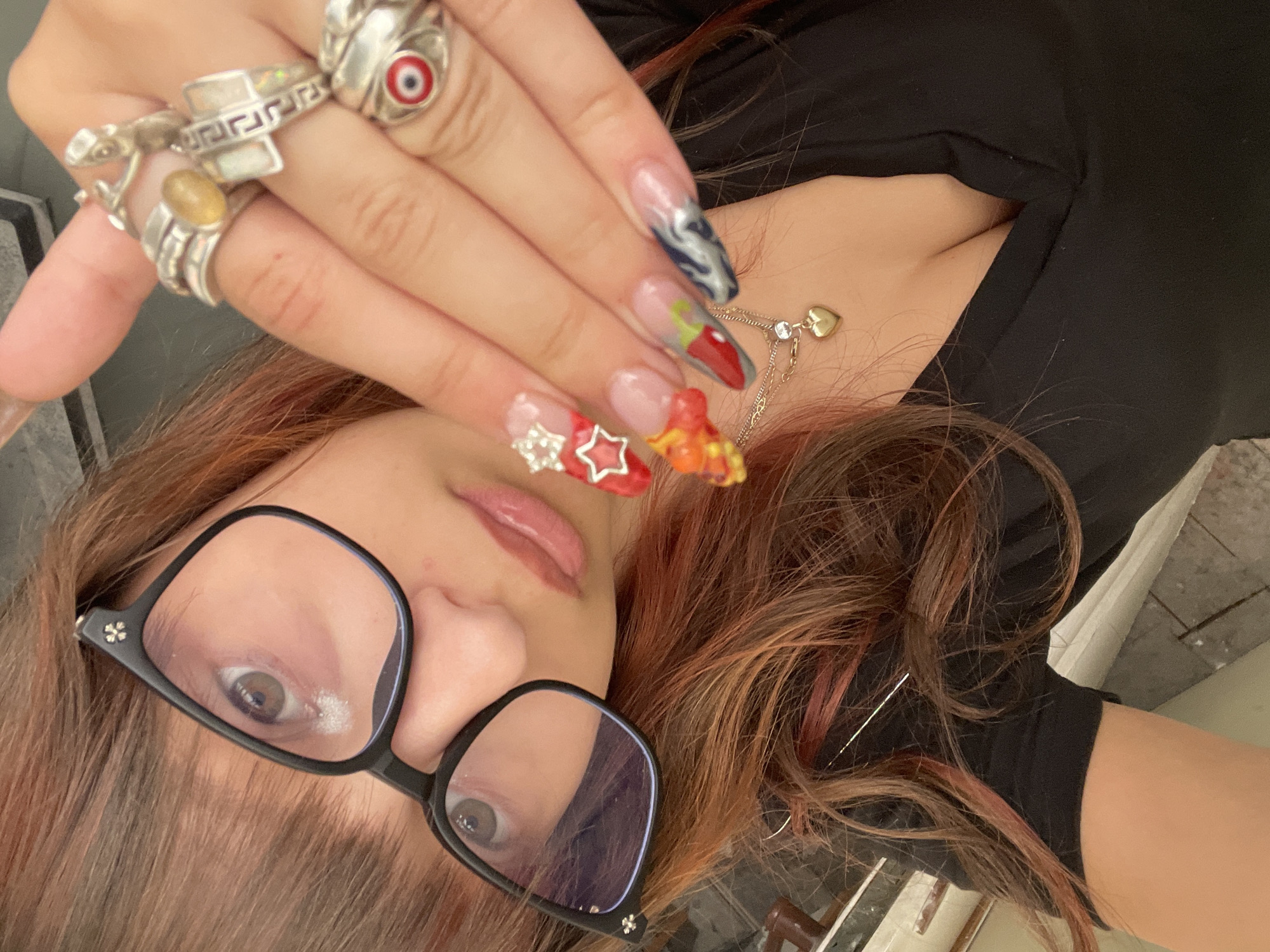 A close-up selfie shows a young woman with reddish-brown hair, wearing black-rimmed glasses and a low-cut black top. She has elaborate acrylic nails, with designs including stars, flames, a chili pepper, and abstract patterns. She is wearing several rings on her right hand, including one with a red evil eye, and a necklace with a gold heart pendant. The background is out of focus but appears to be an indoor setting, possibly a room in a residential building, with some textured flooring visible. The image does not contain any information that would allow for a definitive determination of its location within the city of Raanana, Israel.