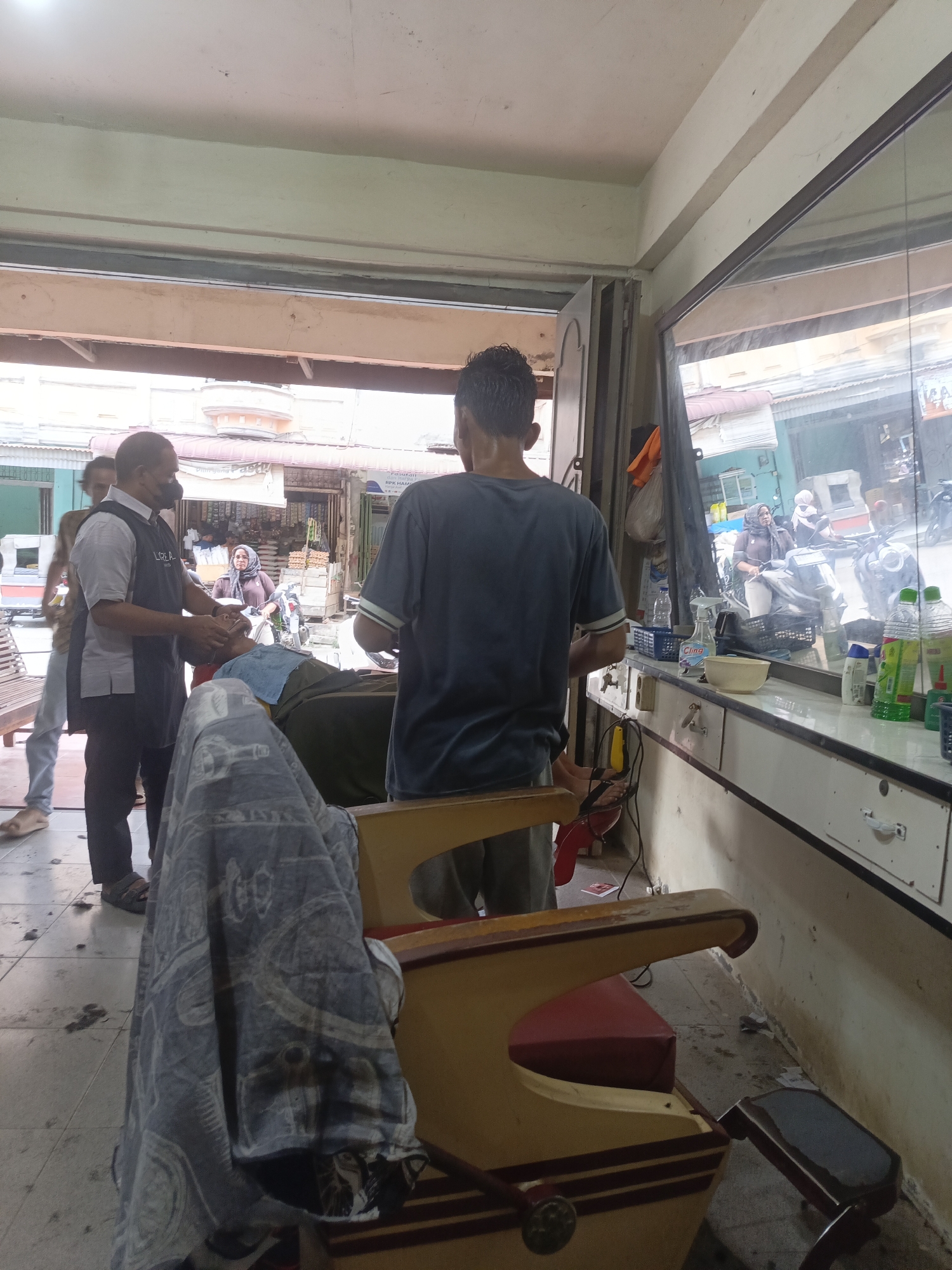 The image shows a barbershop in Lhokseumawe, Indonesia. A barber, wearing a mask and a vest with "L'Oréal" printed on it, is giving a haircut to a client who is lying on a barber chair. Another person is standing to the left, and a man in a blue t-shirt is standing in the foreground, facing away from the camera. The barbershop appears to be an open-air establishment, with a street visible through the entrance. The street is lined with shops and there are motorcycles parked outside. The interior of the barbershop is cluttered with barber tools, bottles of hair products, and a mirror that reflects the street scene. The floor is dusty and there are some hair clippings on the ground. The overall impression is of a busy, working barbershop in a bustling urban environment.